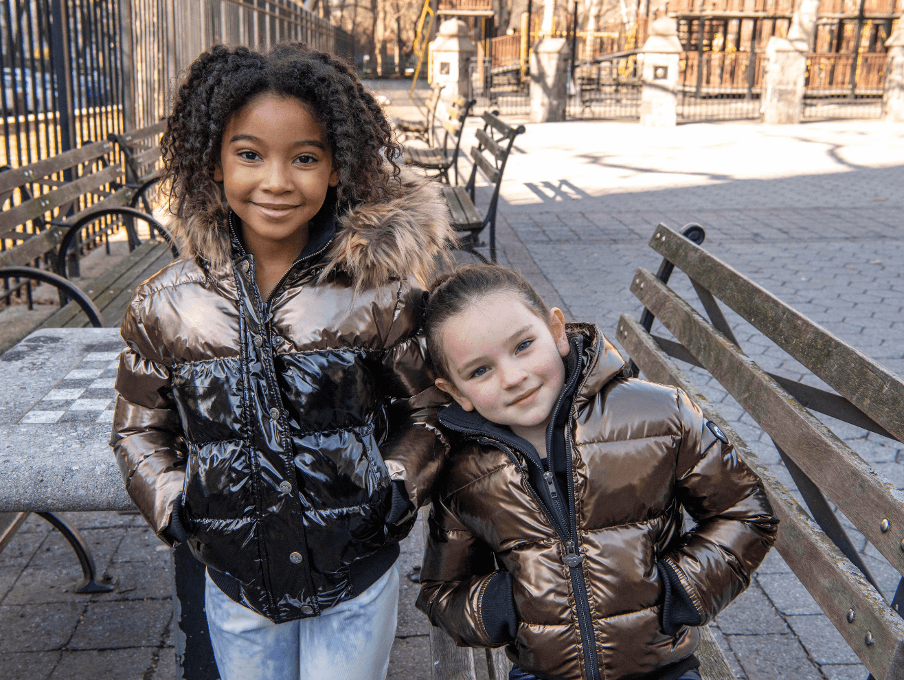 Two stylish kids in shiny puffy jackets enjoying a sunny day in the park, showcasing playful winter fashion.
