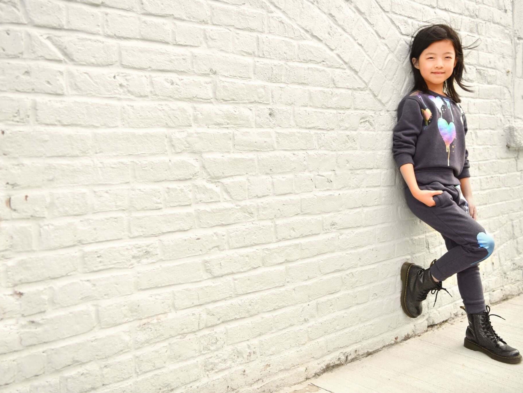 Stylish girl in playful Appaman outfit, showcasing trendy kids' fashion against a chic white brick wall.