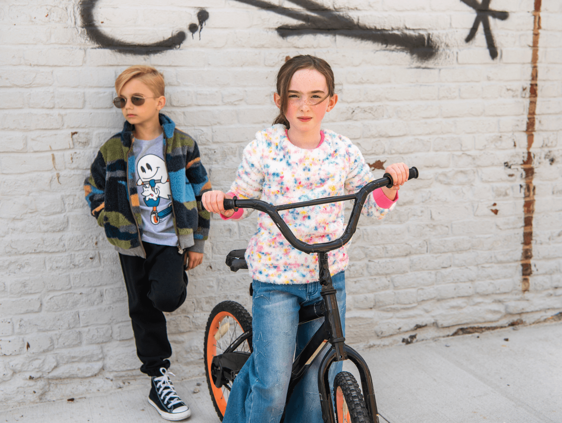 Two stylish kids, one on a bike and the other with sunglasses, showcasing trendy back-to-school outfits in a graffiti setting.
