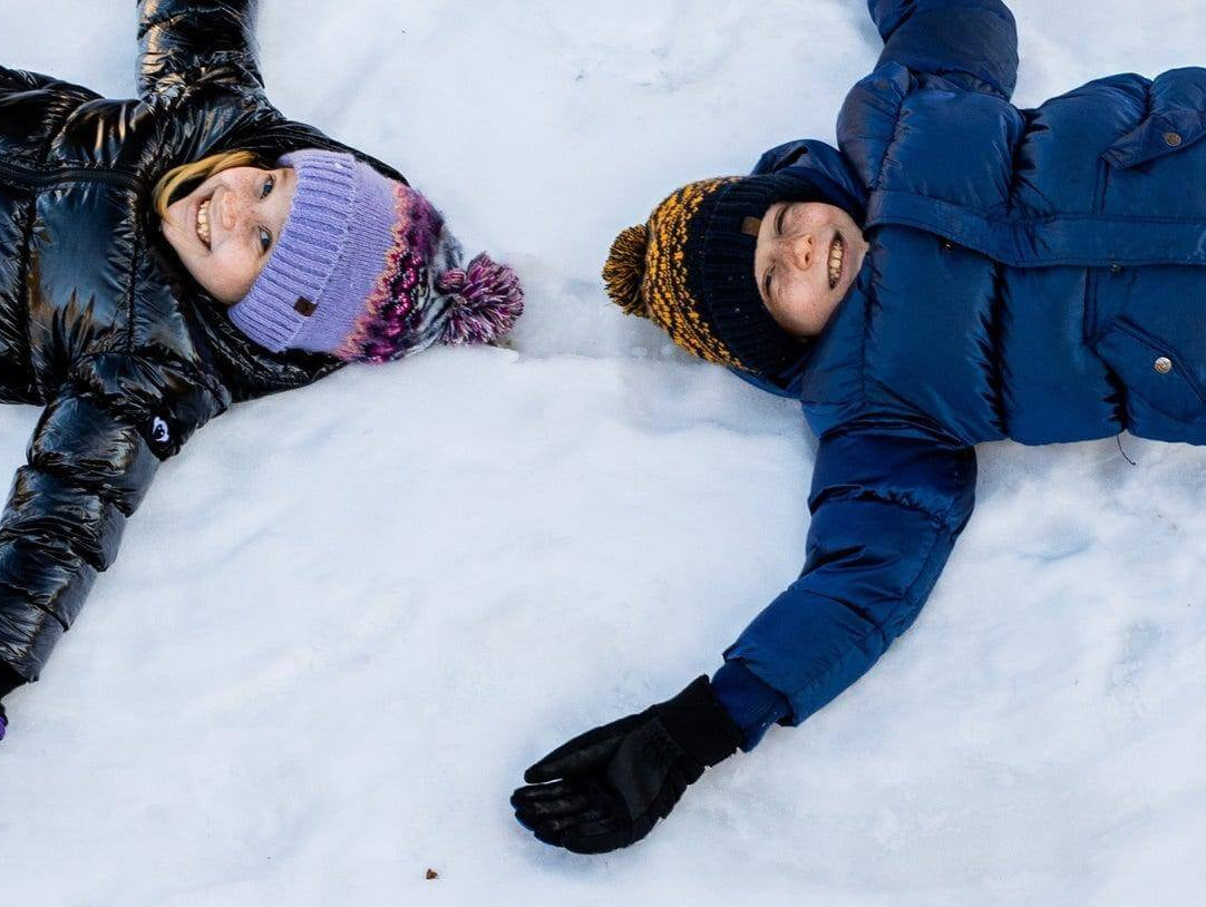 Two kids joyfully making snow angels in a winter wonderland, bundled up in stylish jackets and gloves, embracing the season's fun.
