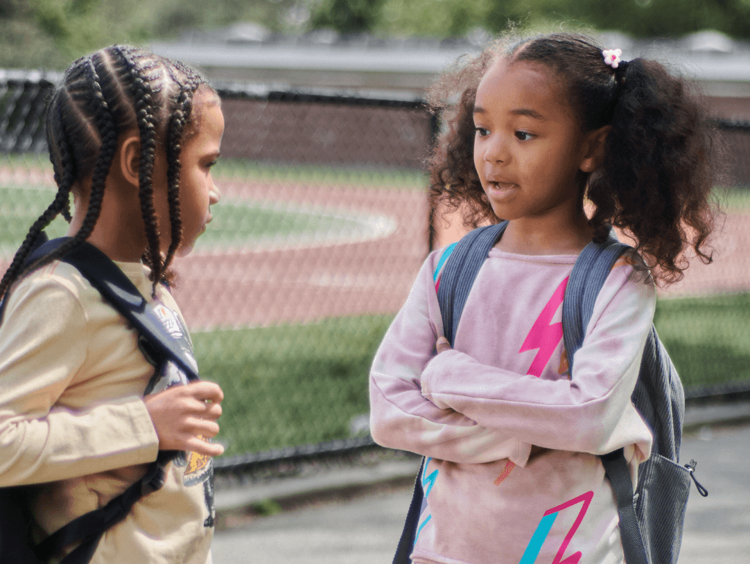 Two stylish girls chatting outdoors, showcasing trendy outfits and backpacks for school.