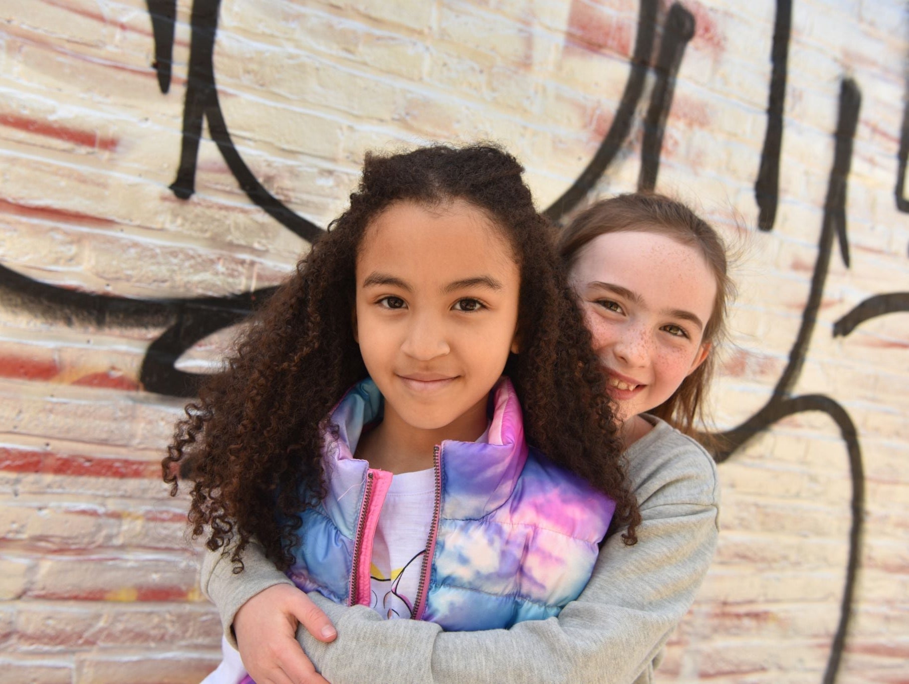Two stylish girls sharing a joyful moment, rocking colorful Appaman outfits against a vibrant graffiti wall.