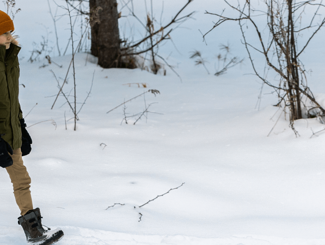 A stylish kid in a cozy green jacket and orange hat walks through snowy woods, ready for family winter fun.