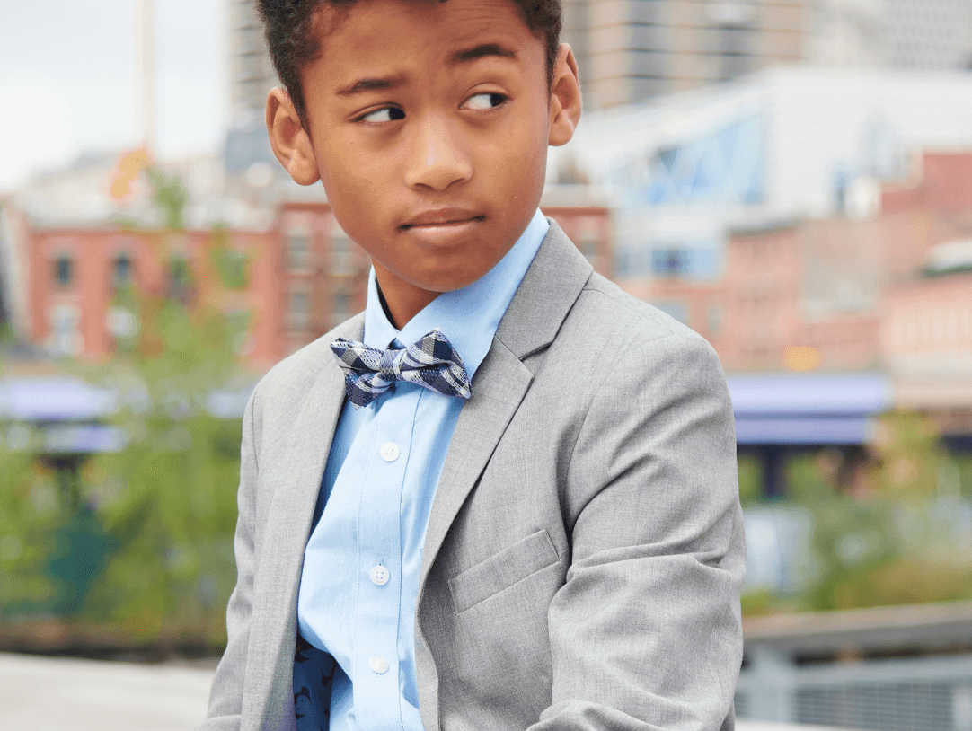 Dapper young boy in a gray suit and bow tie, showcasing stylish kids fashion in a vibrant urban setting.