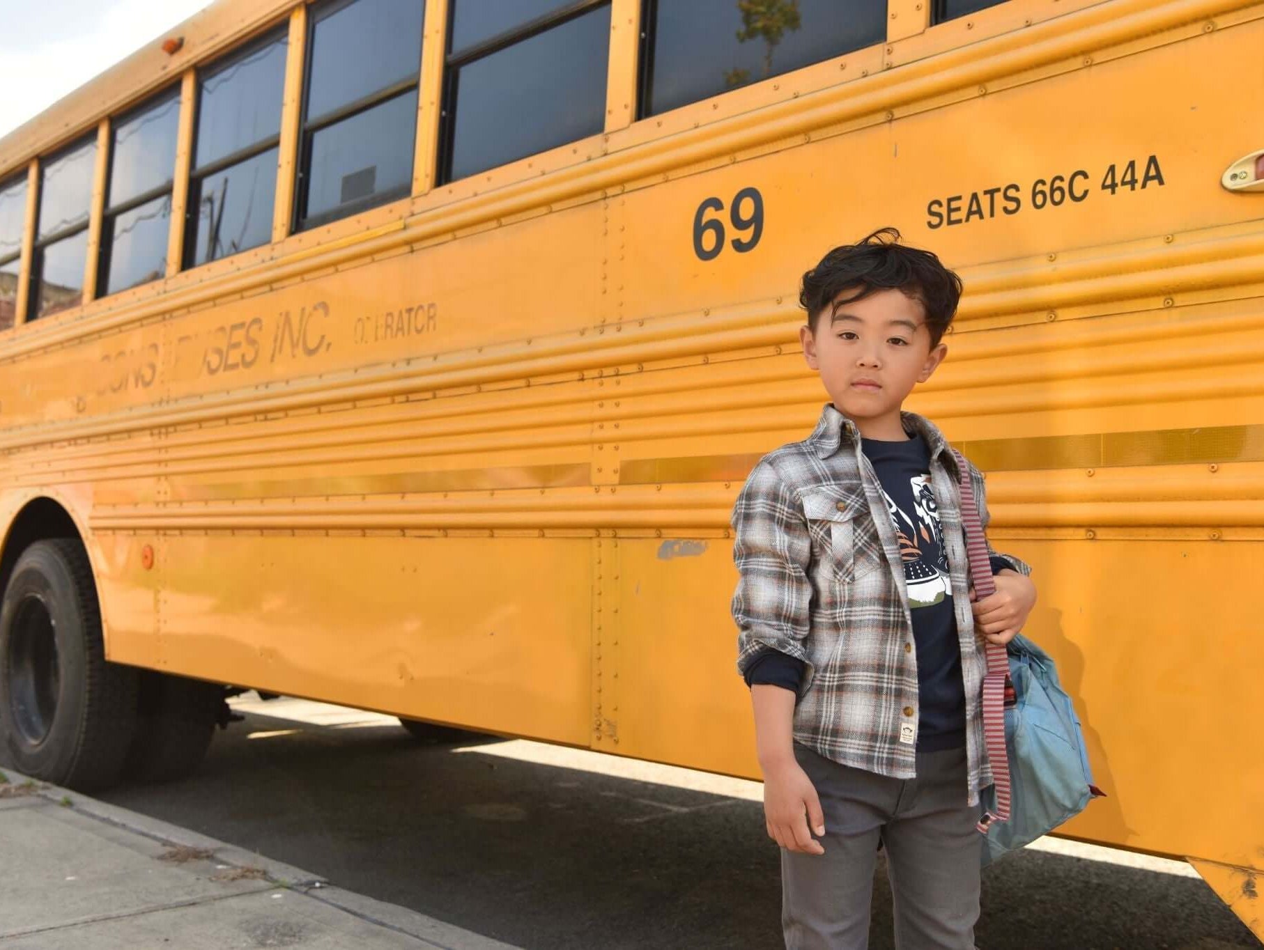 Stylish boy in a plaid jacket stands next to a bright yellow school bus, showcasing trendy kids' outfit ideas.