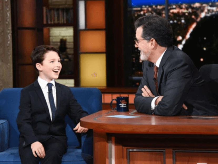 Young boy in a suit joyfully chatting with Stephen Colbert on a late-night talk show, showcasing stylish kids' fashion.