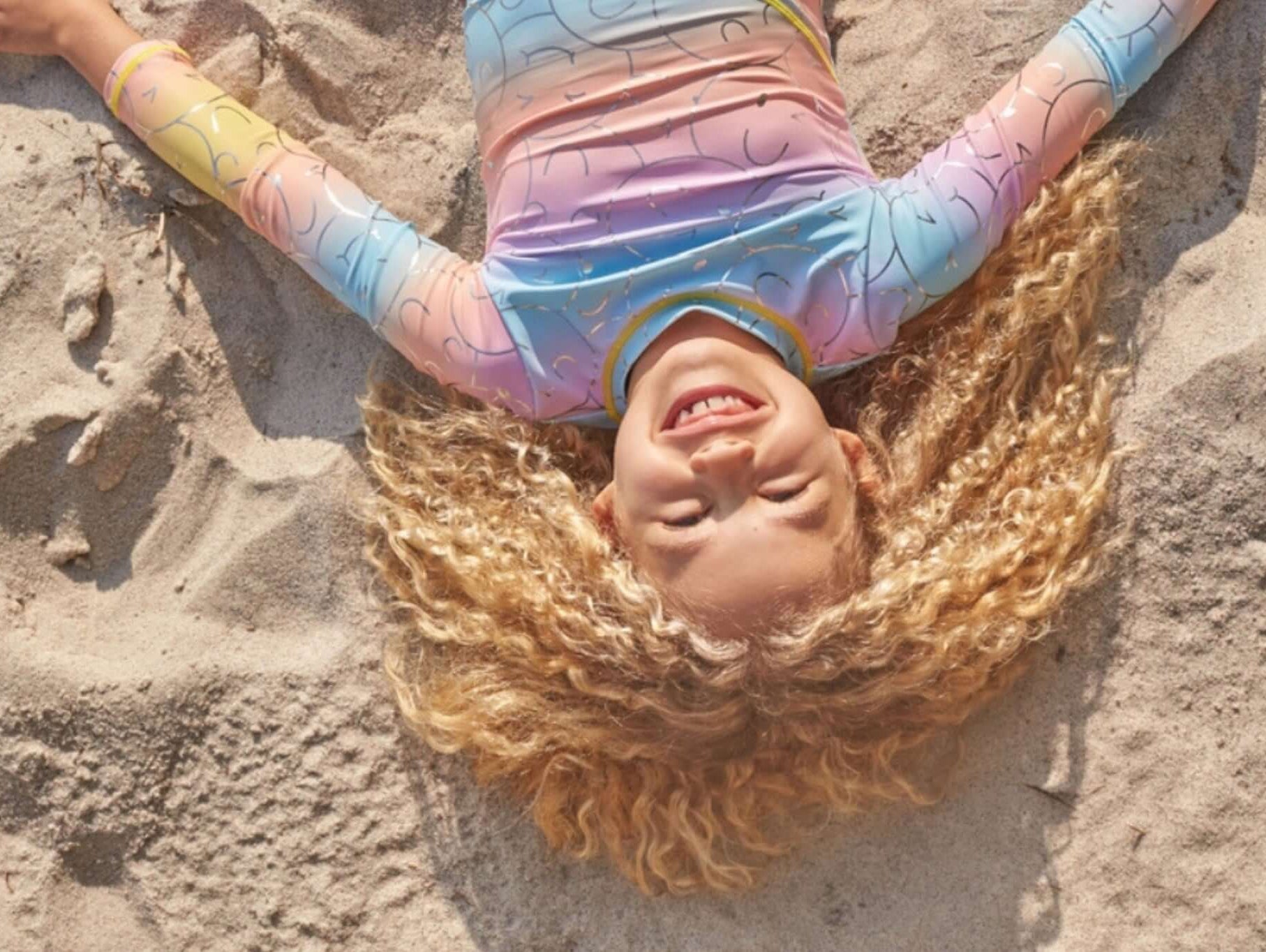 A joyful girl with curly hair smiles widely while lying on the sandy beach, showcasing her colorful swimsuit.