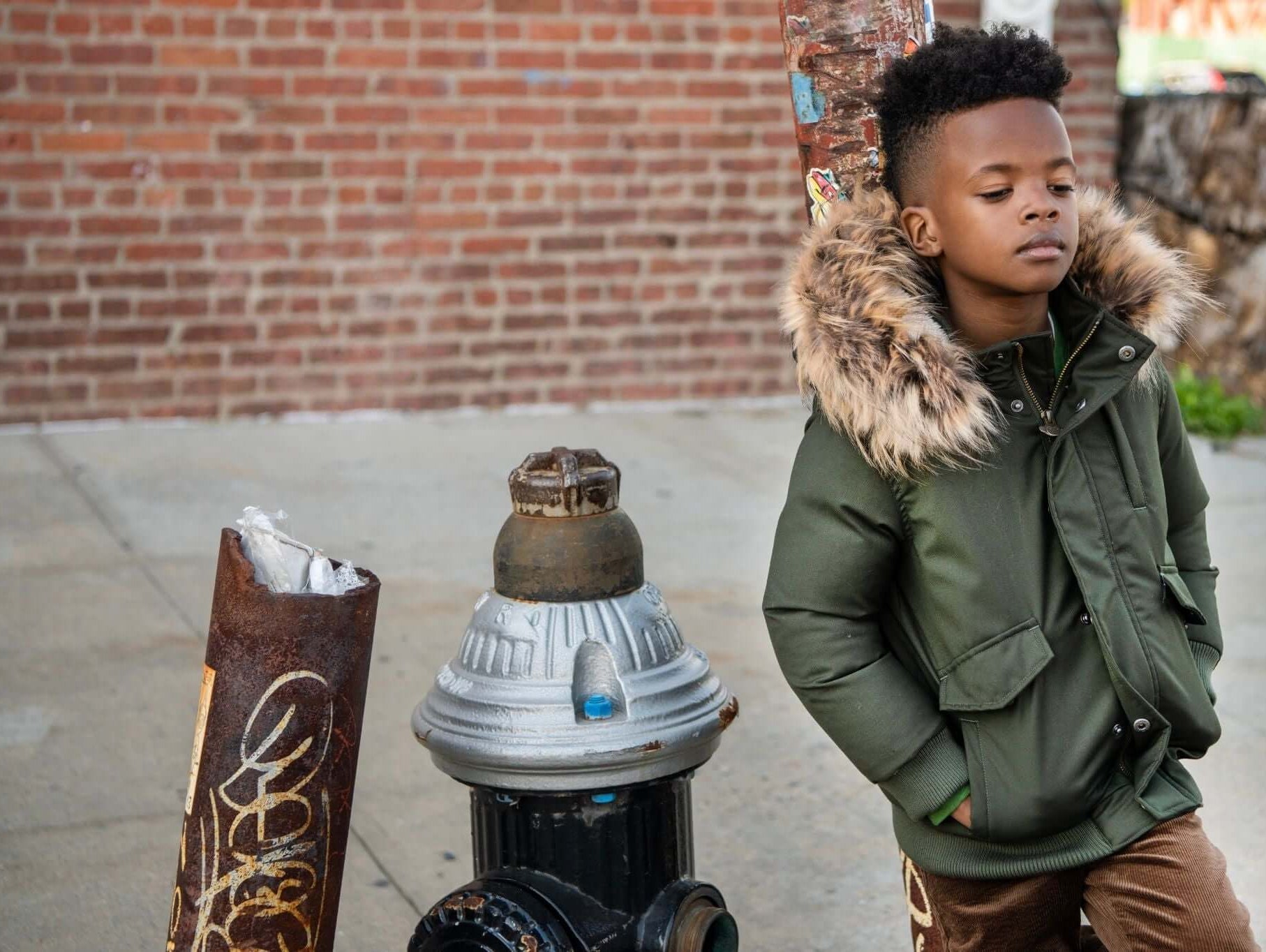 Stylish boy in a green down jacket with fur trim, posing confidently by a graffiti-covered fire hydrant.