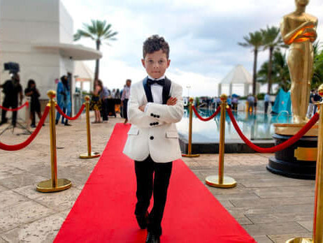 Stylish young boy in a tuxedo walks confidently on the red carpet, ready to steal the spotlight at a glamorous event.