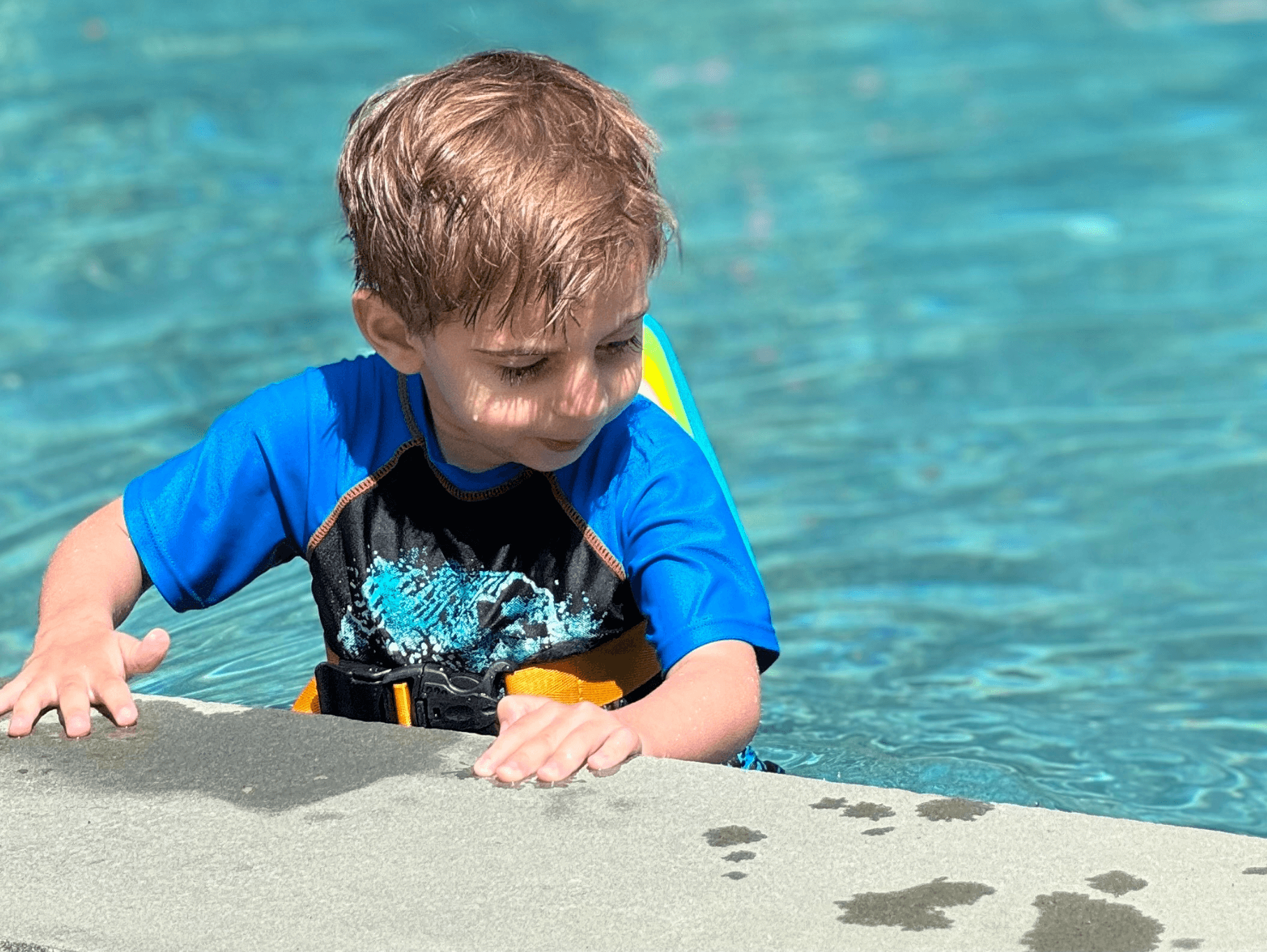 A young boy in a stylish swim shirt joyfully splashes at the edge of a sparkling pool, ready for summer fun.