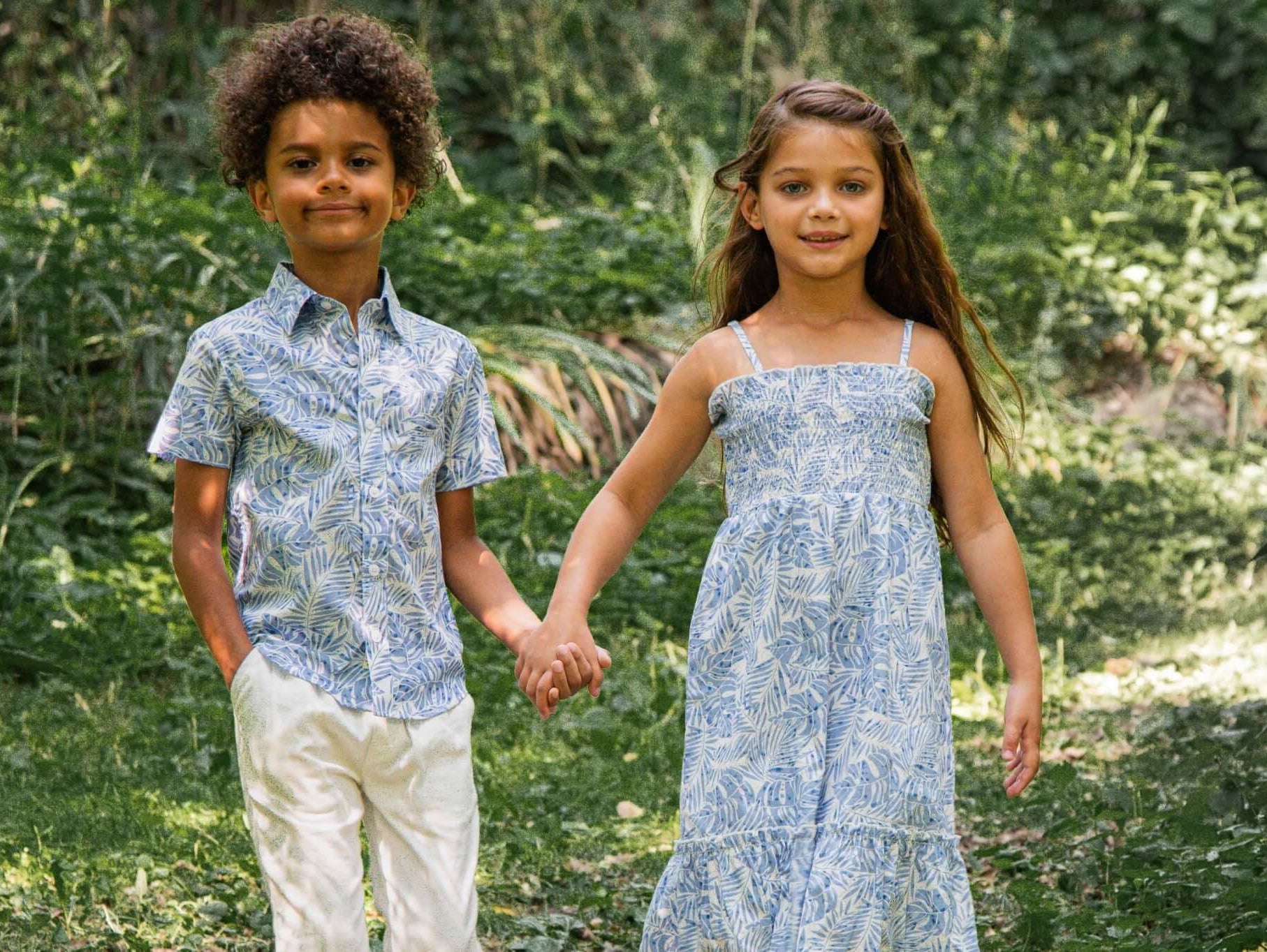 Stylish kids' summer outfits: a boy in a floral print shirt and white pants, and a girl in a blue dress, holding hands in nature.