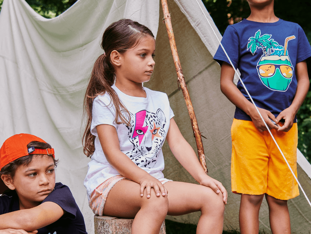 Kids having fun outdoors in stylish summer camp outfits by a teepee, showcasing playful and trendy summer styles.