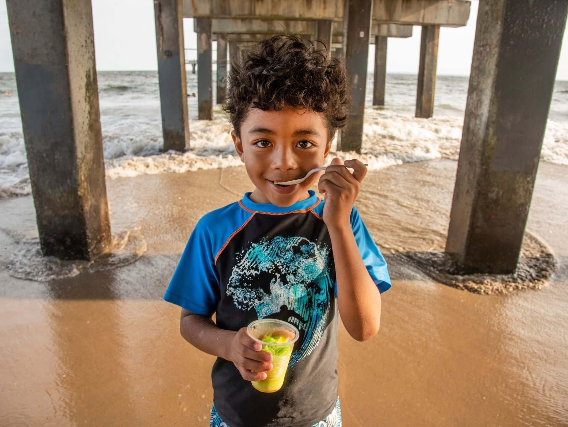 Smiling boy enjoying a refreshing treat under a pier, wearing colorful swimwear, perfect for summer family fun!