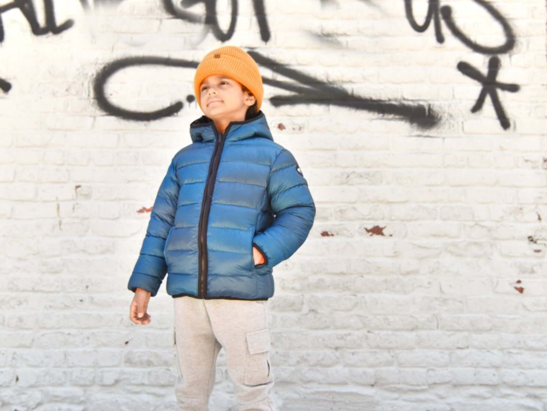 Stylish boy in a blue puffer jacket and orange beanie, showcasing trendy kids' winter fashion against a graffiti wall.