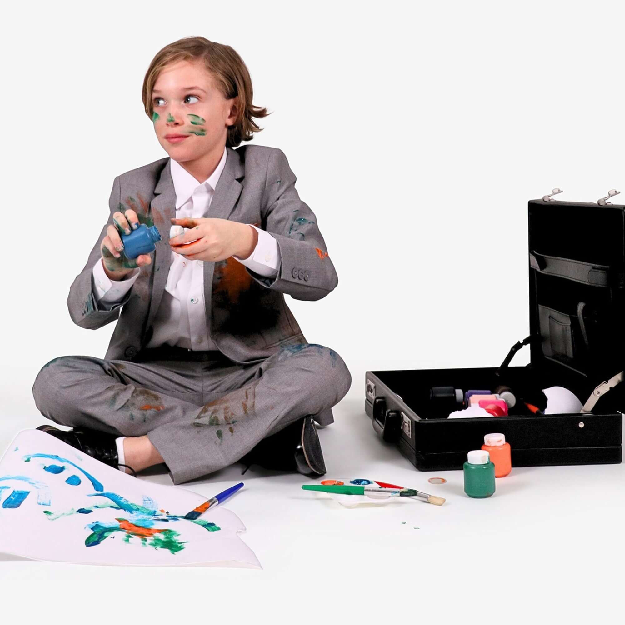 Stylish kid in a washable suit, having fun creating art with paint and a suitcase of craft supplies.