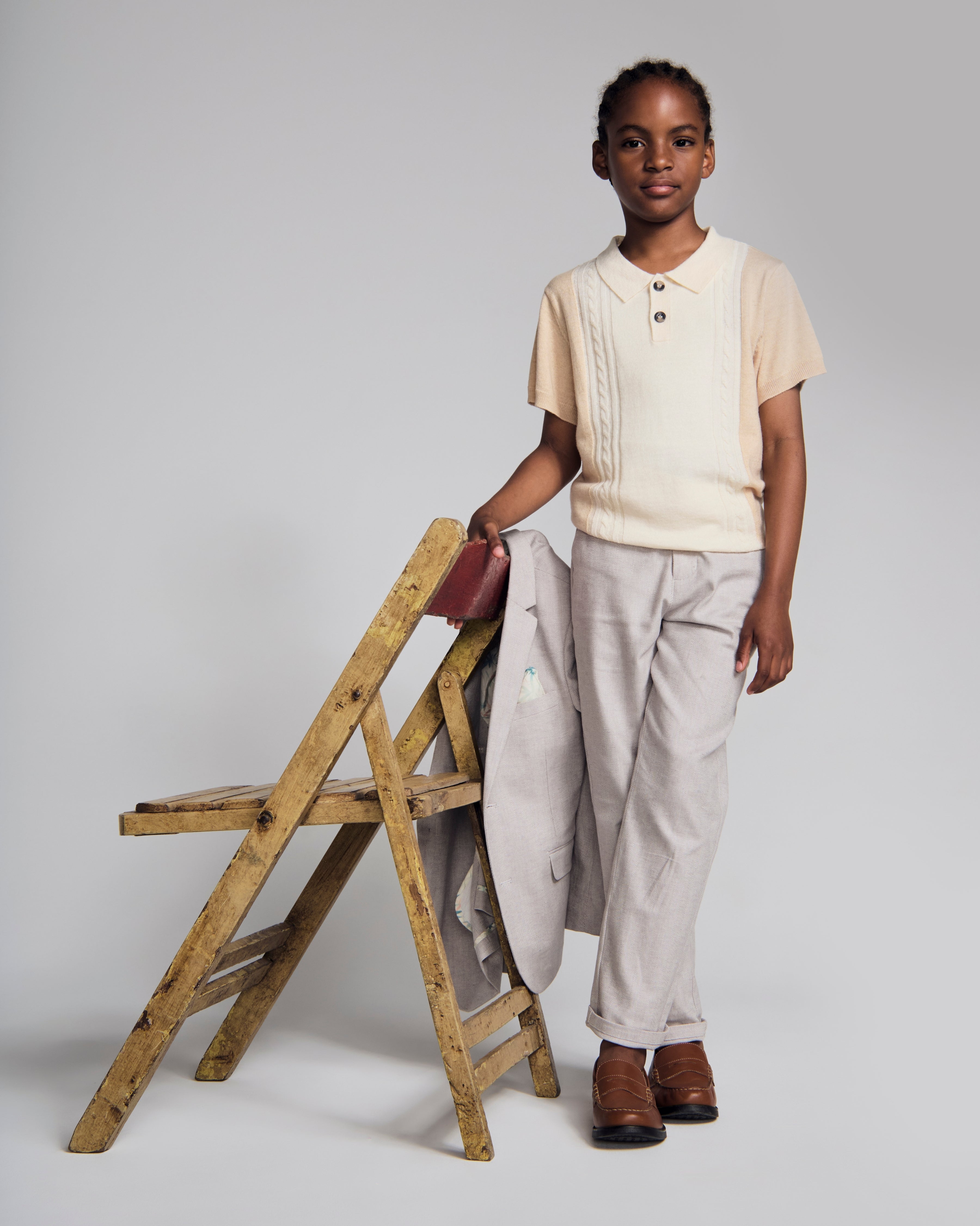 A child wearing a Beige Sweater Knit Polo with collared neckline, paired with light gray pants, standing by a wooden chair.