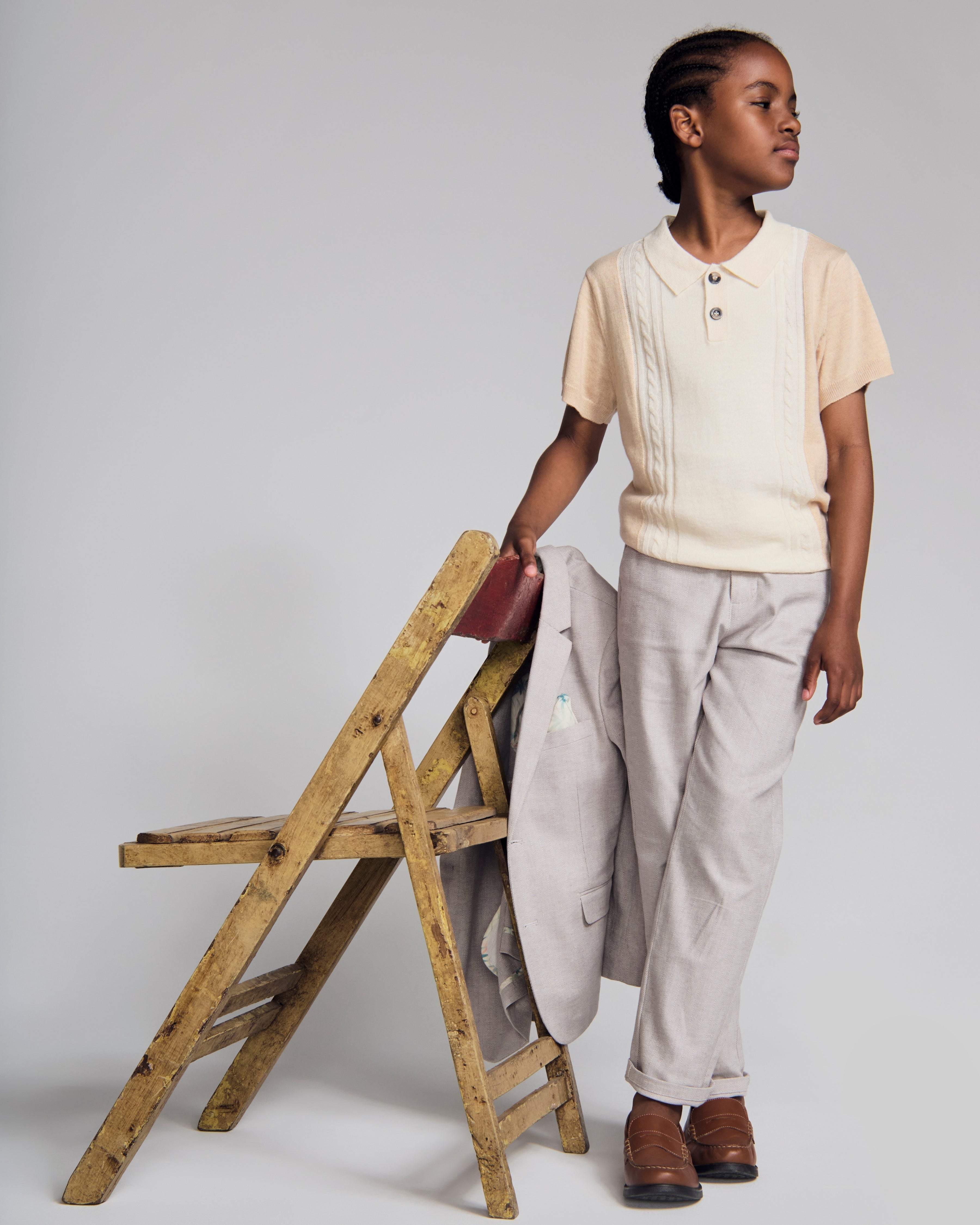 Boy wearing a Beige Sweater Knit Polo with light grey trousers, standing beside a wooden chair.