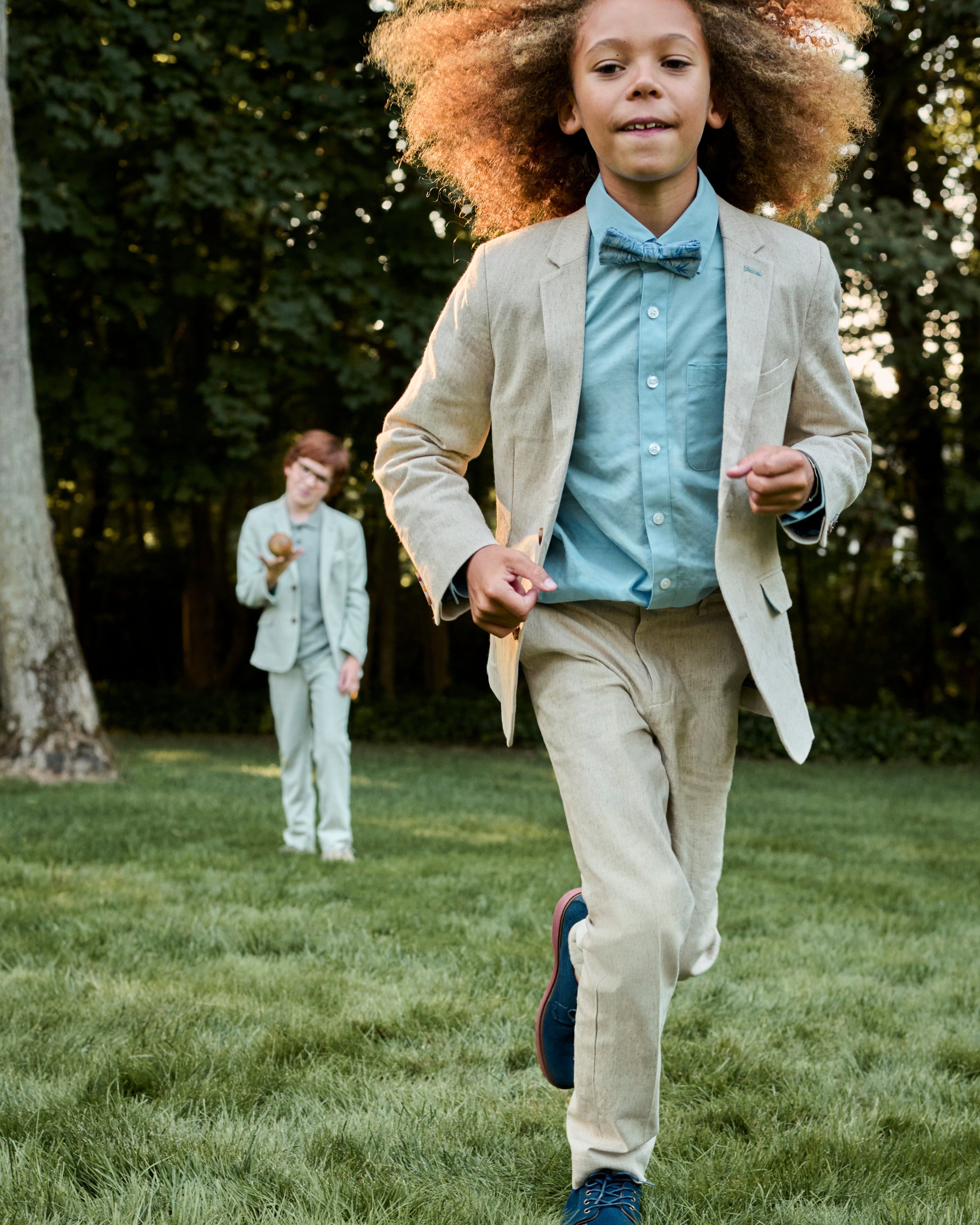 Two children in formal attire running outdoors on a grassy area with trees in the background.