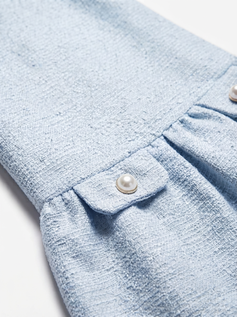 Close-up of a light blue textured fabric garment detail, showing a pleated section, a small flap pocket with a pearl button, and part of a matching pearl button on the sleeve.