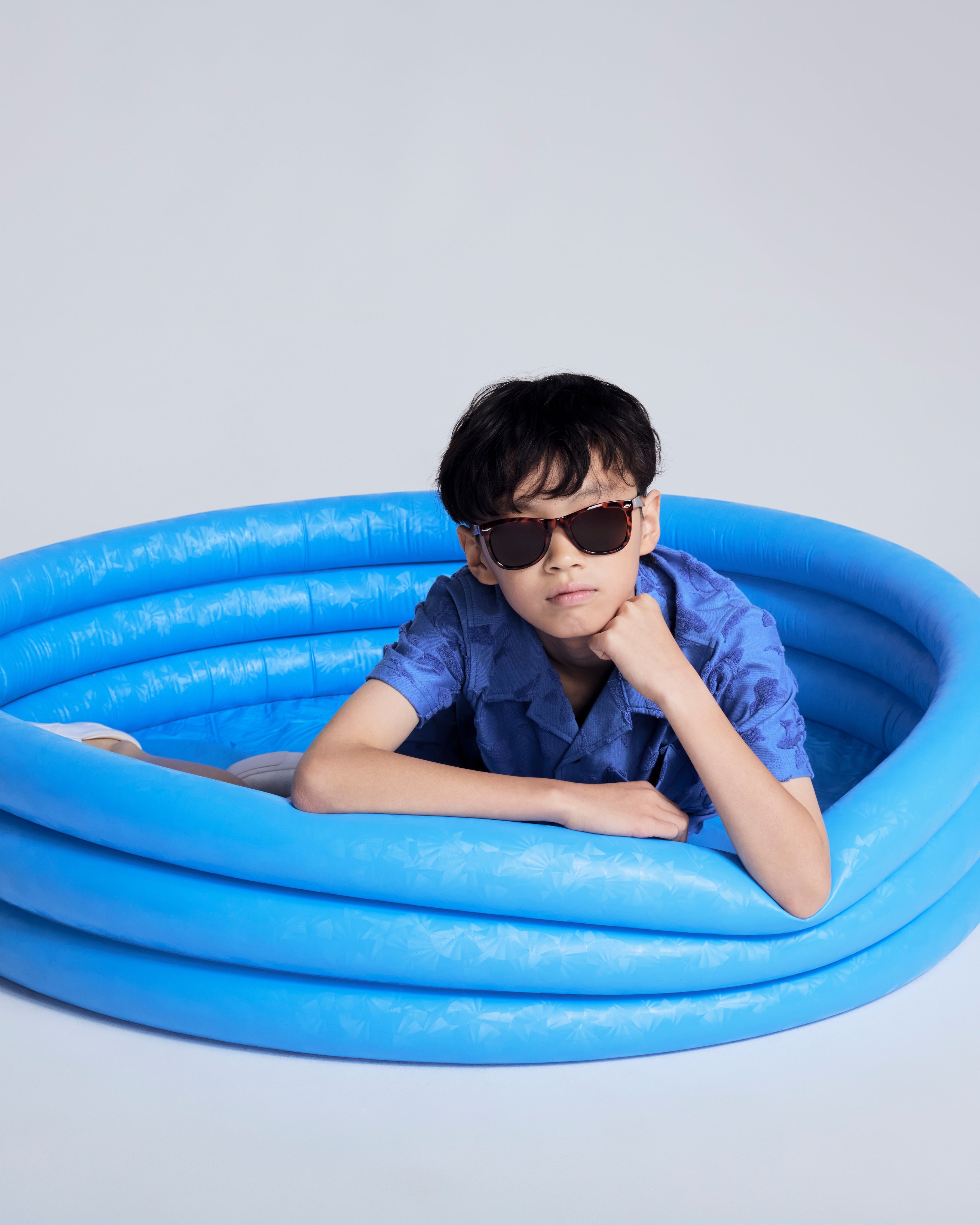 Boy wearing Cobalt Blue Terry Resort Shirt lounging in a blue inflatable pool with sunglasses.