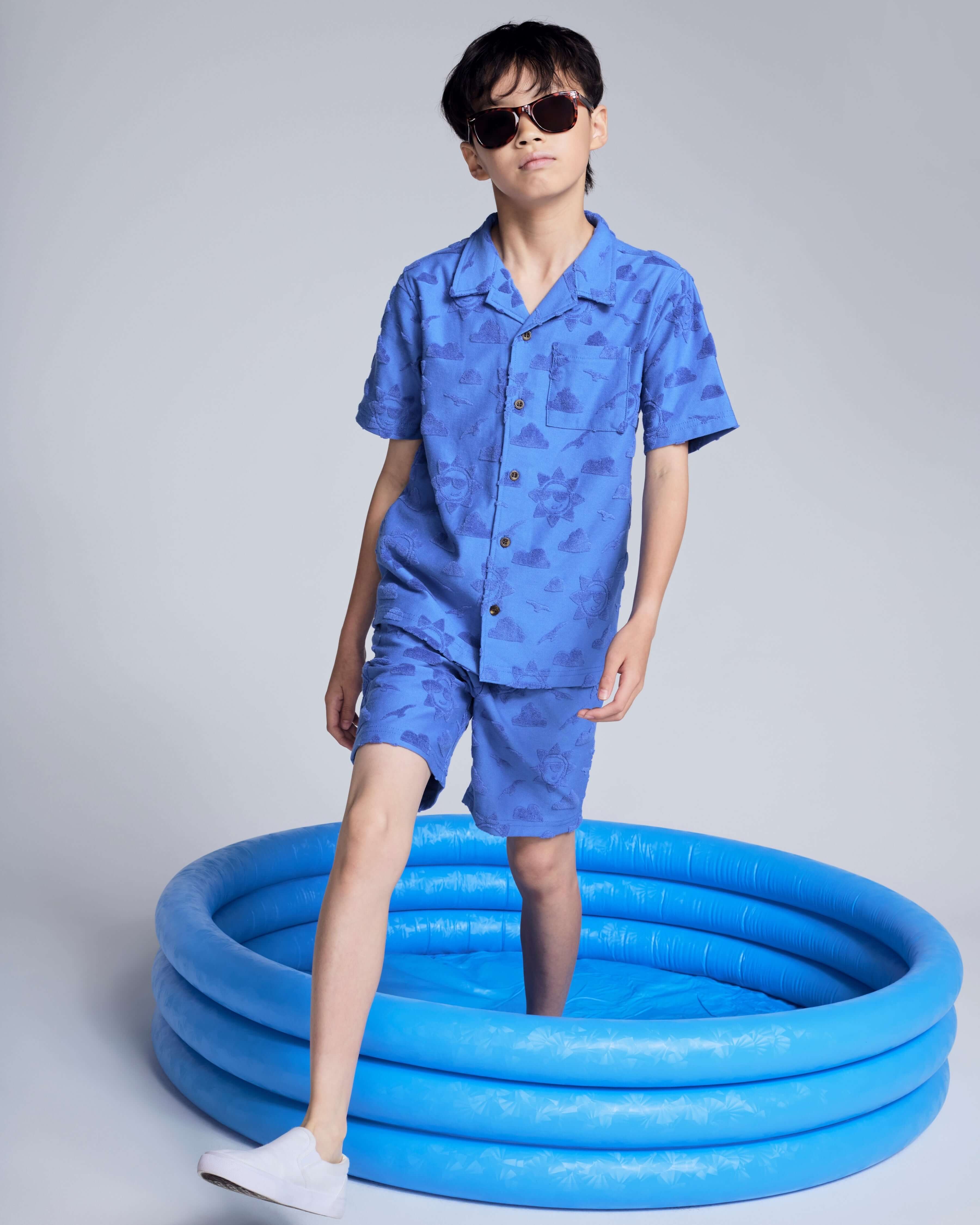 Boy wearing Cobalt Blue Terry Resort Short and matching shirt, sunglasses, standing in a blue inflatable pool.