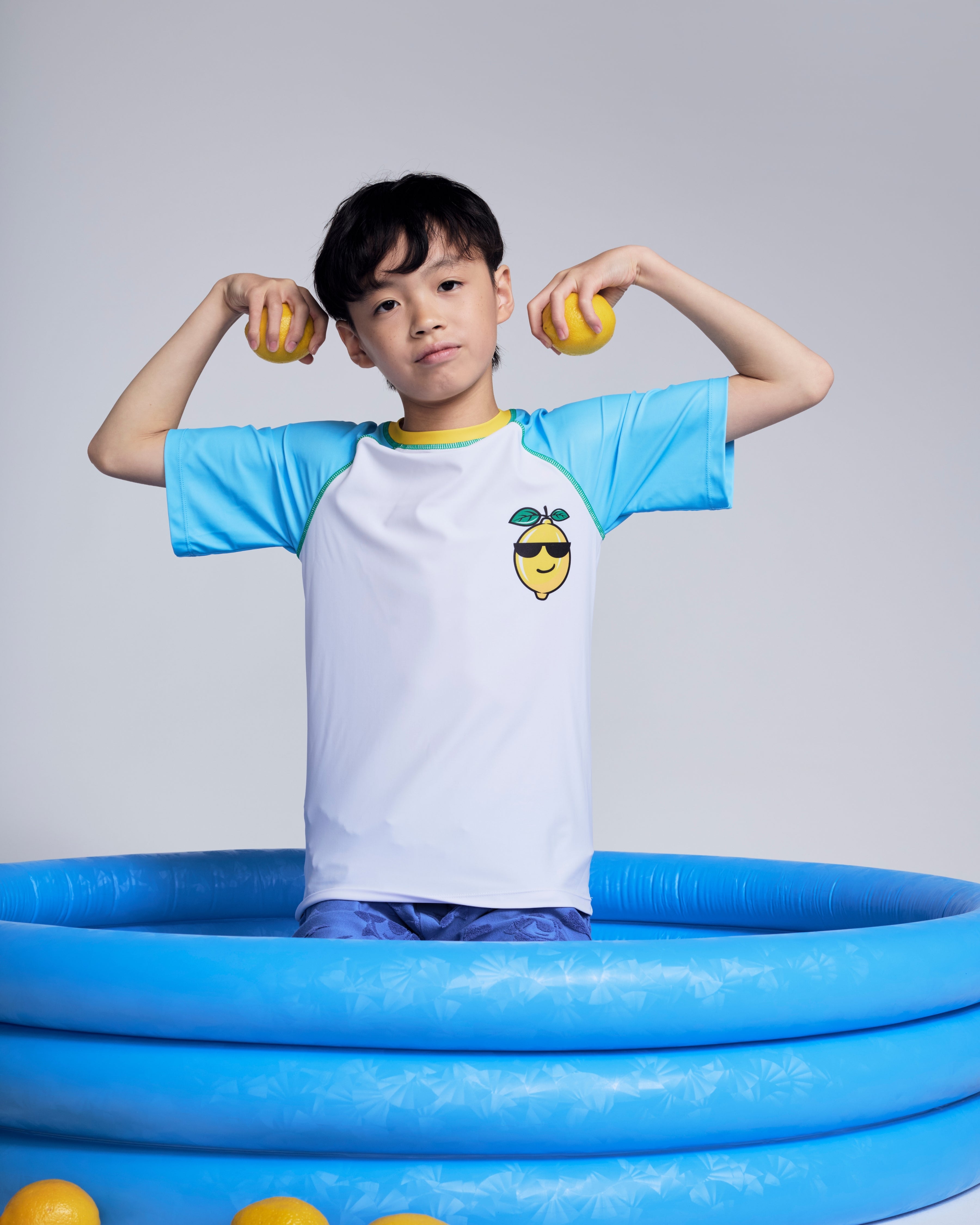 Boy wearing a Cool Lemon Rash Guard in a blue pool holding lemons, showcasing summer fun and protection.