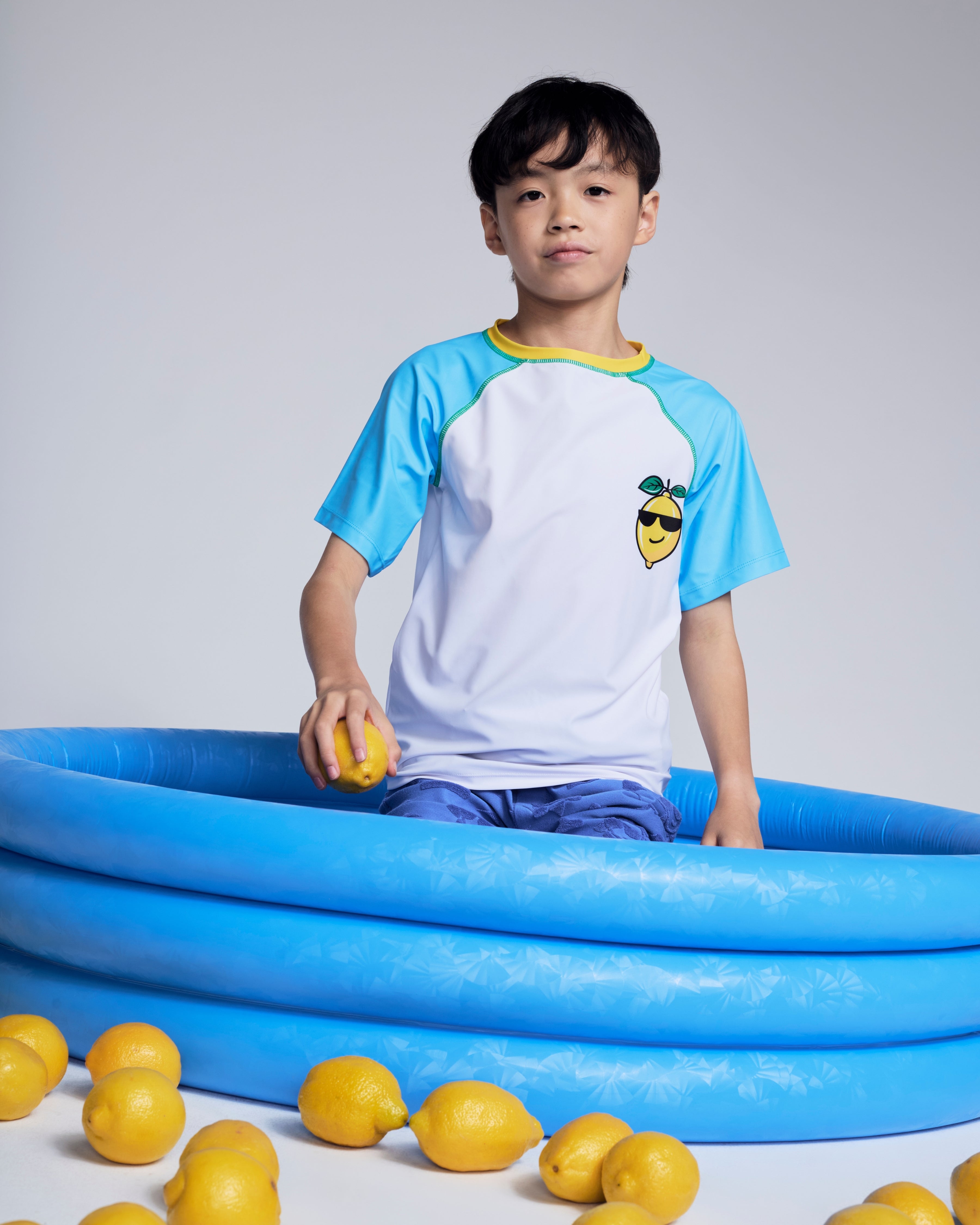 Boy wearing a Cool Lemon Rash Guard sitting in a blue inflatable pool surrounded by lemons.