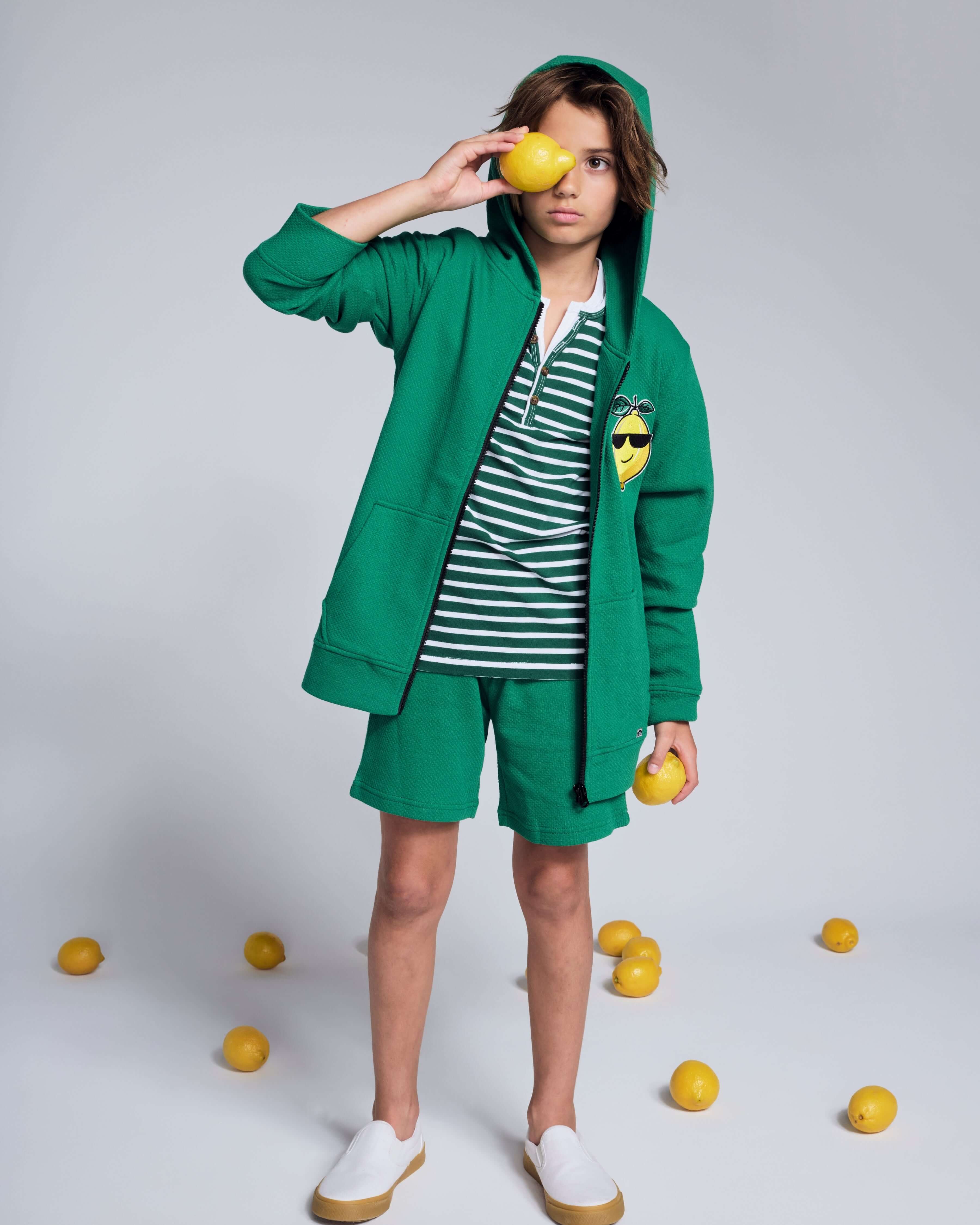 Boy wearing Garden Green Resort Short and hooded jacket, holding lemons in summer play setting.