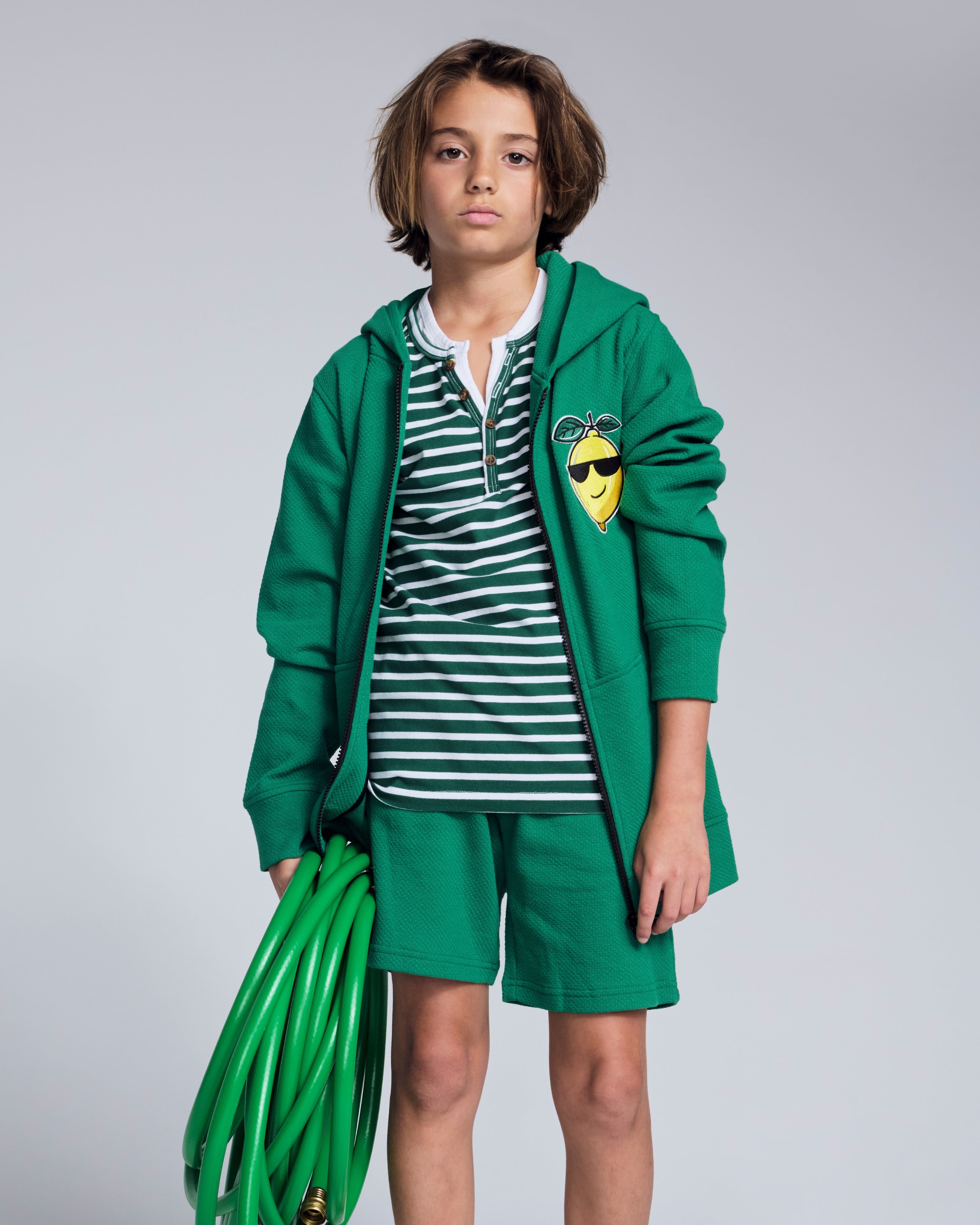 Child wearing a green hoodie and Garden Green Stripe Hilltop Henley with a garden hose.