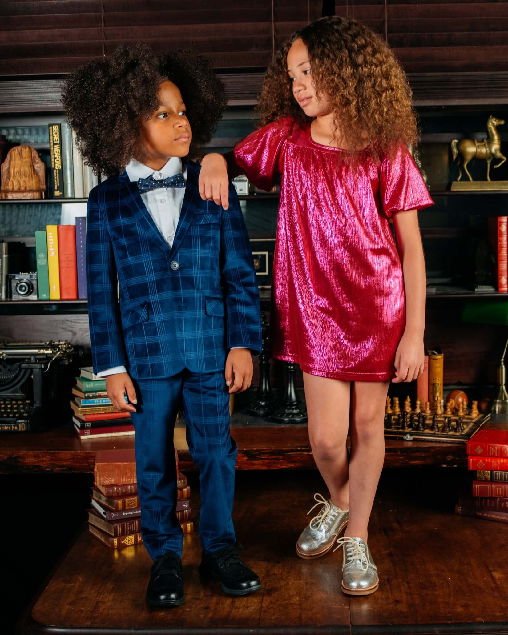 Two children posing in a stylish setting, featuring a boy in a blue suit and a girl in a Fuchsia Emma Dress.