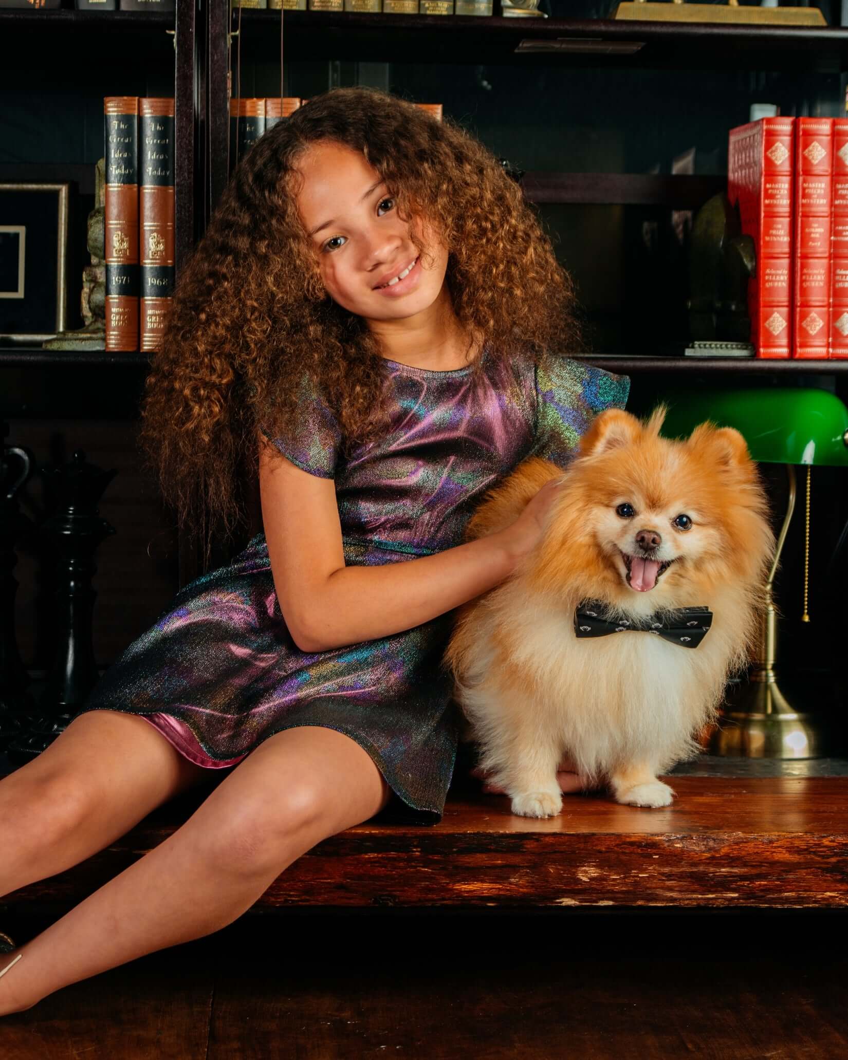 A girl in a Black Rainbow Ellie Dress with flutter cap sleeves poses with a fluffy dog wearing a bow tie in a cozy setting.