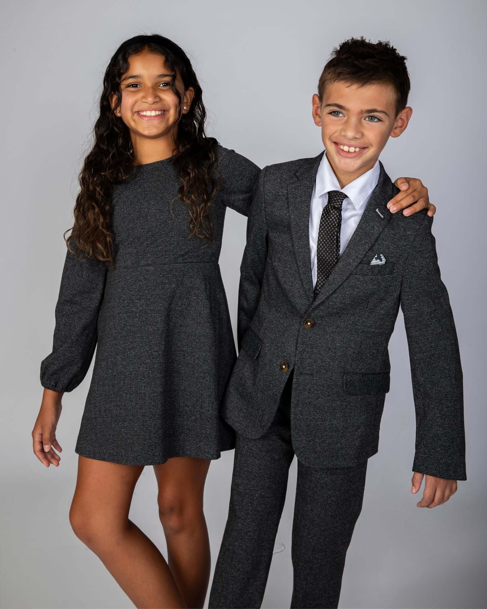 Girl in Noir Micro Plaid Blair Bow Dress with boy in gray suit, both smiling against neutral background.