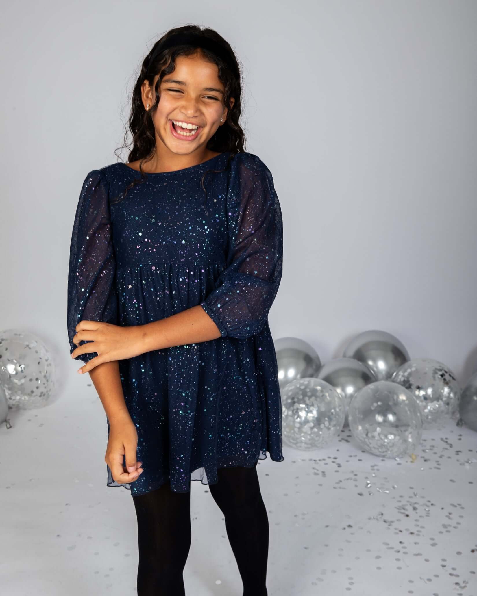 Girl wearing a Navy Blue Chelsea Bow Dress with sparkly fabric, smiling and surrounded by festive decor.
