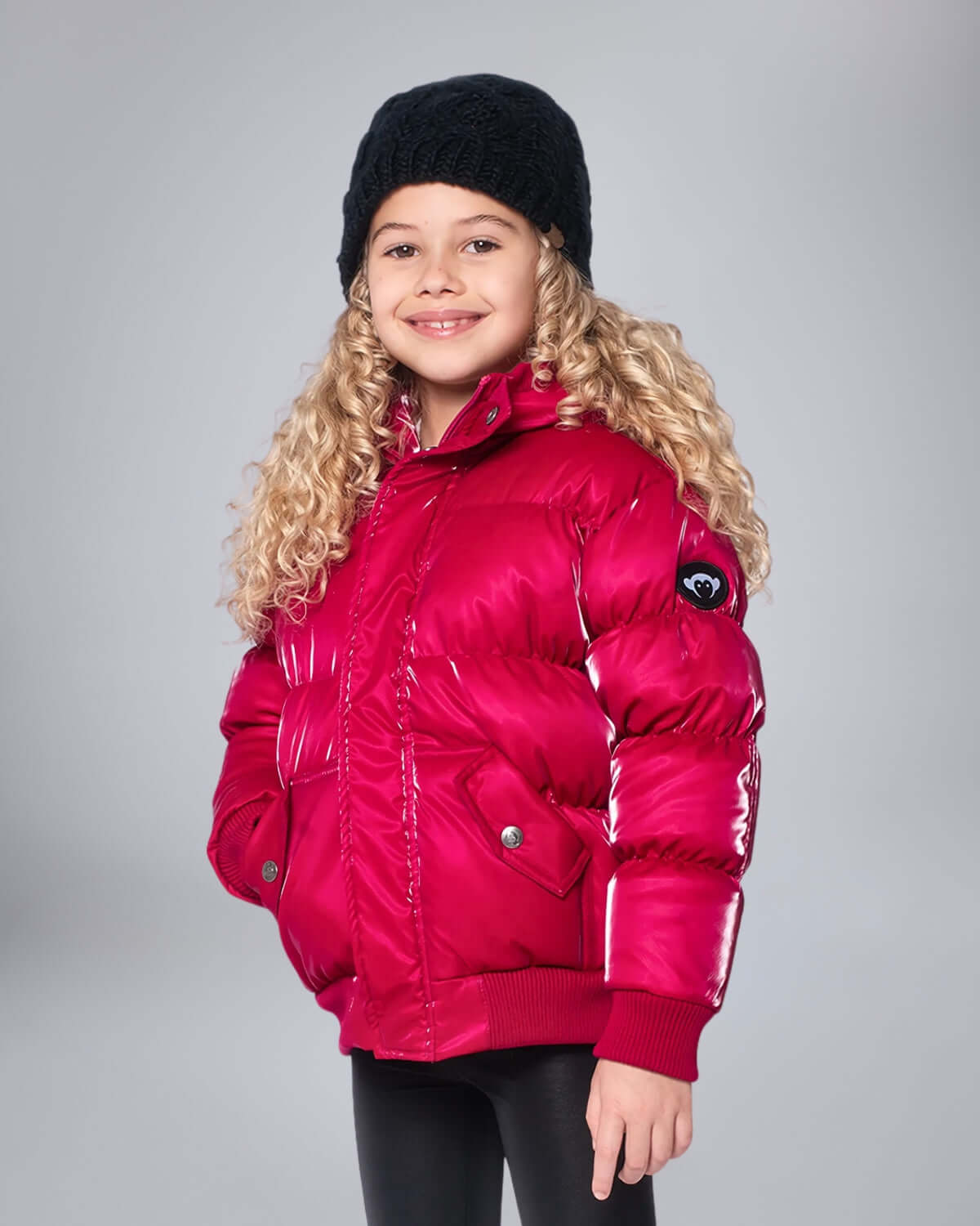 Child wearing a pink puffer jacket and black beanie on a gray background