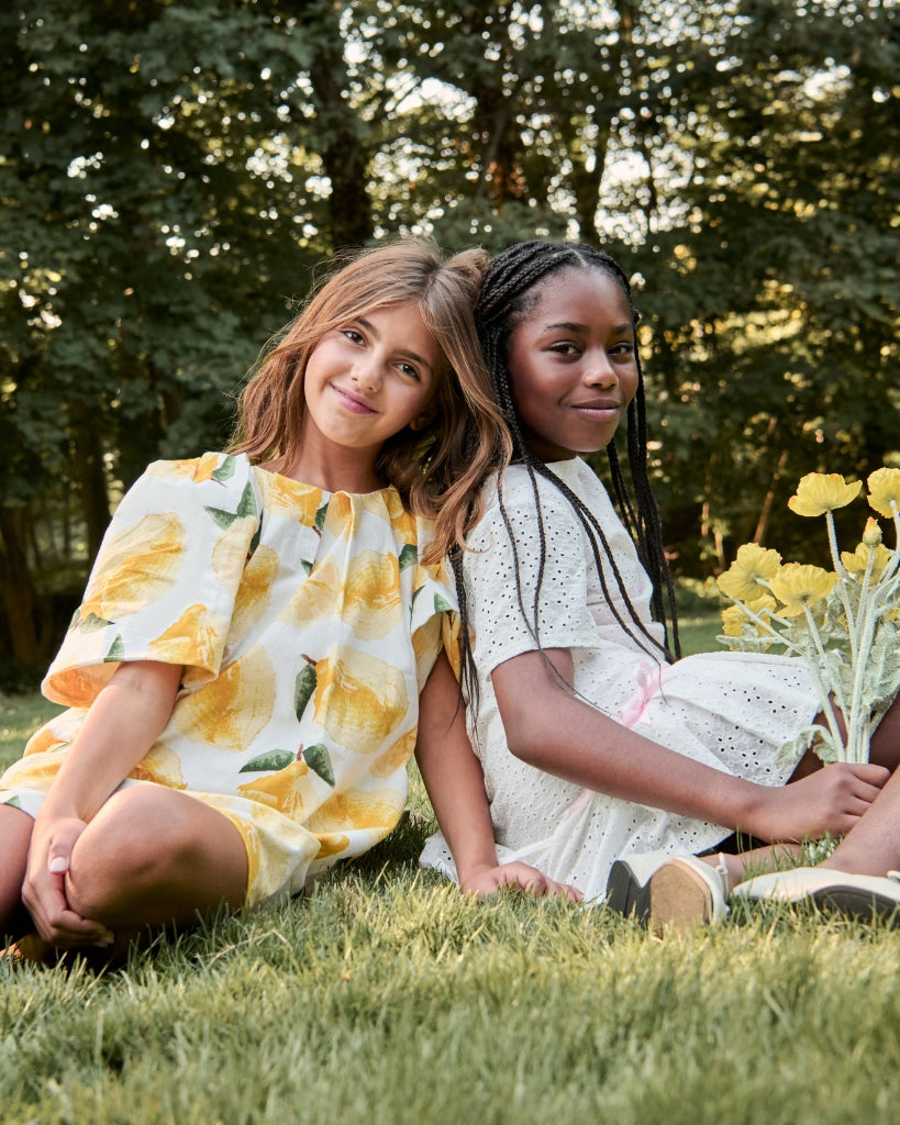 Two young girls sitting on grass holding yellow flowers, surrounded by greenery.