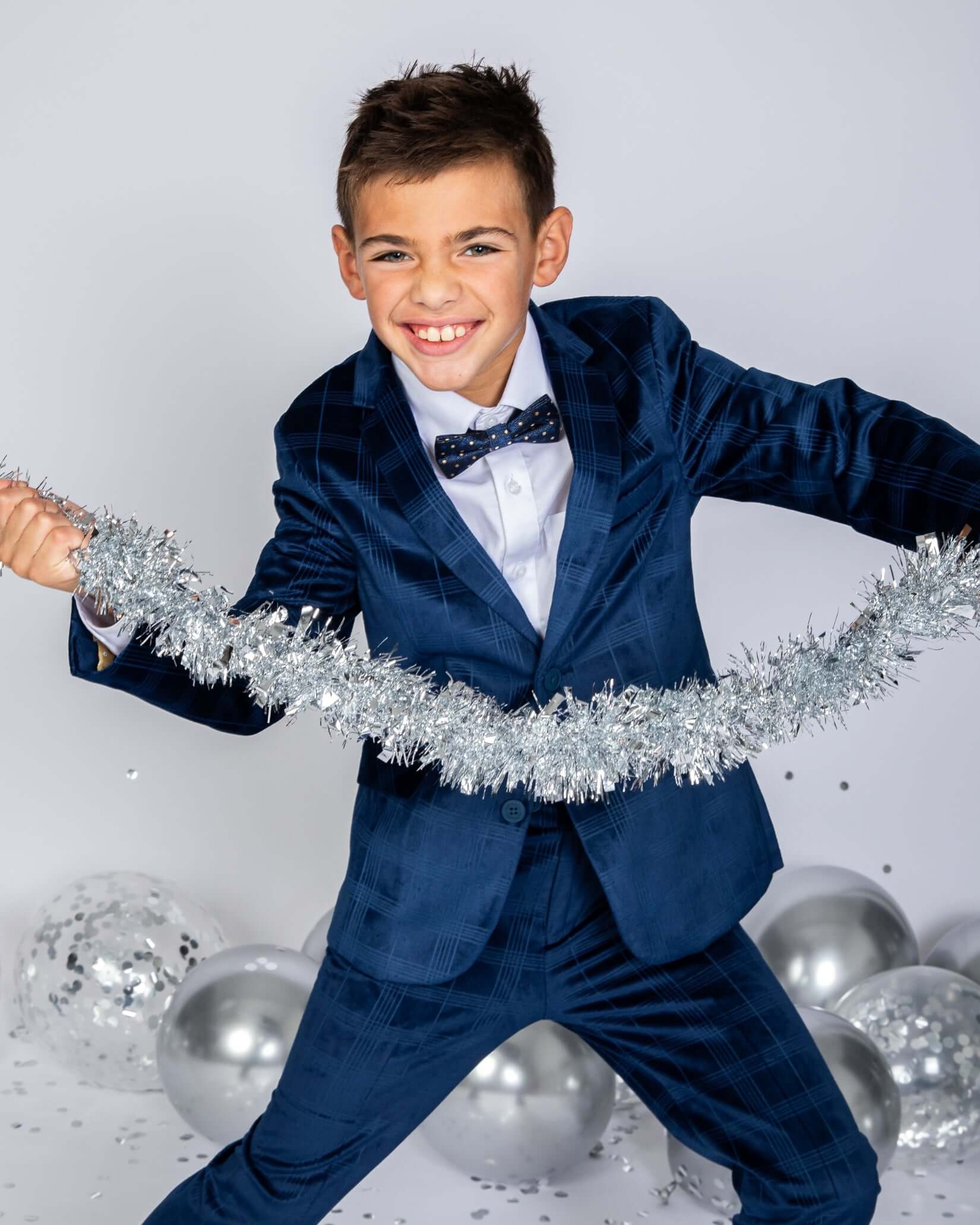 A cheerful boy wearing a Blue Velvet Suit Blazer, holding tinsel and celebrating with silver balloons.