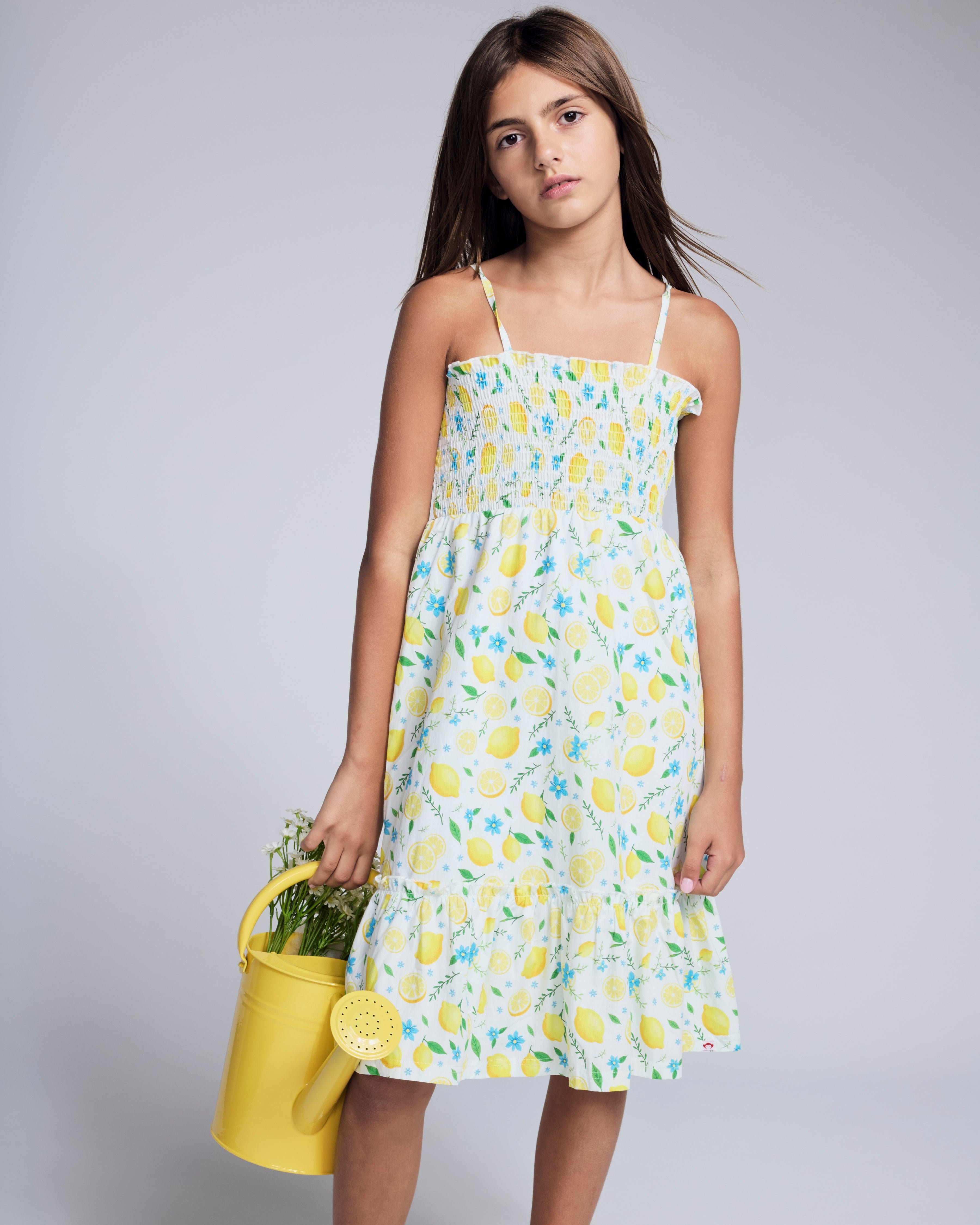 Girl wearing a Lemon Zest Madison Dress with lemon print, holding a yellow watering can, perfect for sunny days.