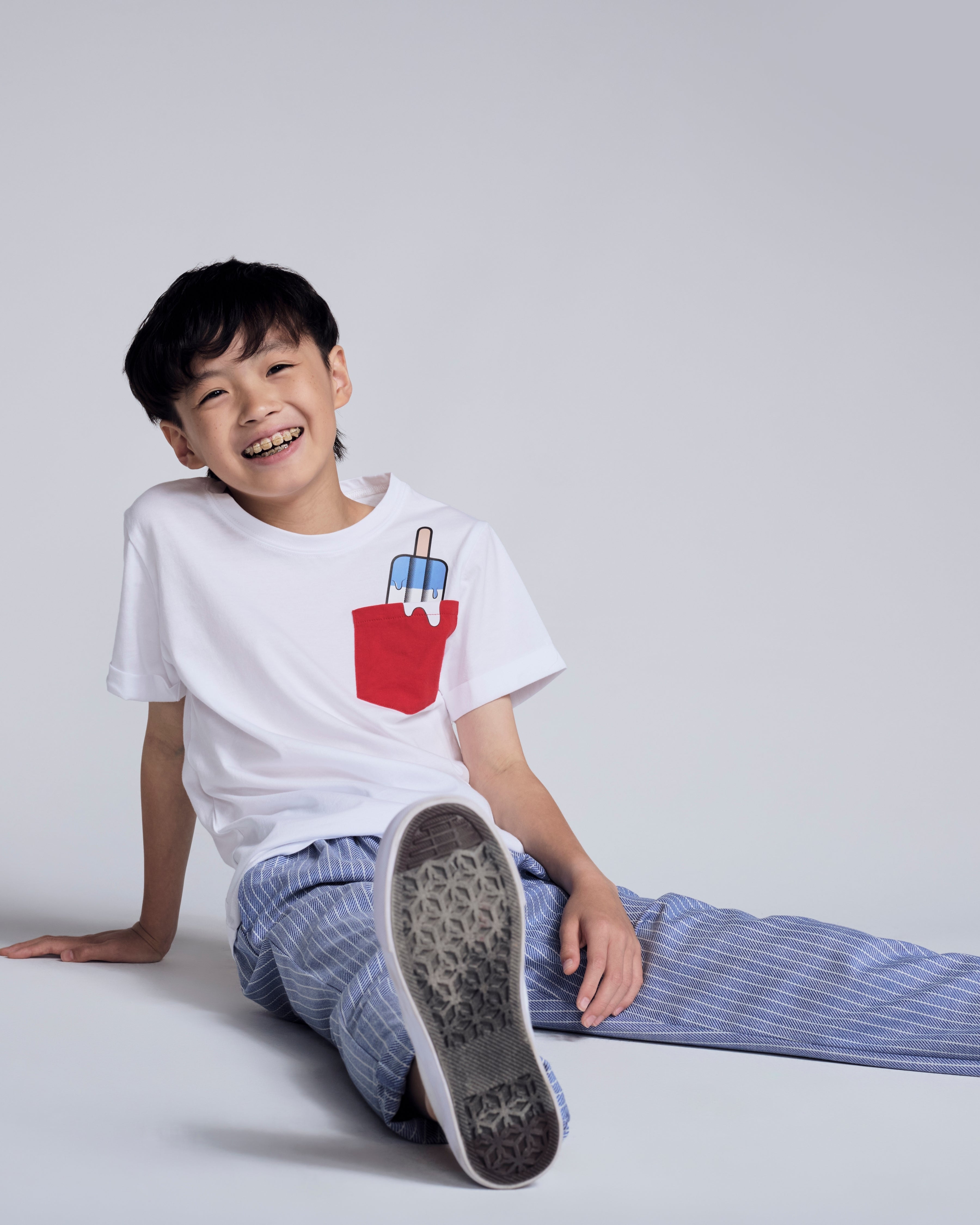 A smiling boy wearing a white shirt with a red pocket and blue striped pants, showcasing the Nautical Stripe Resort Pant.