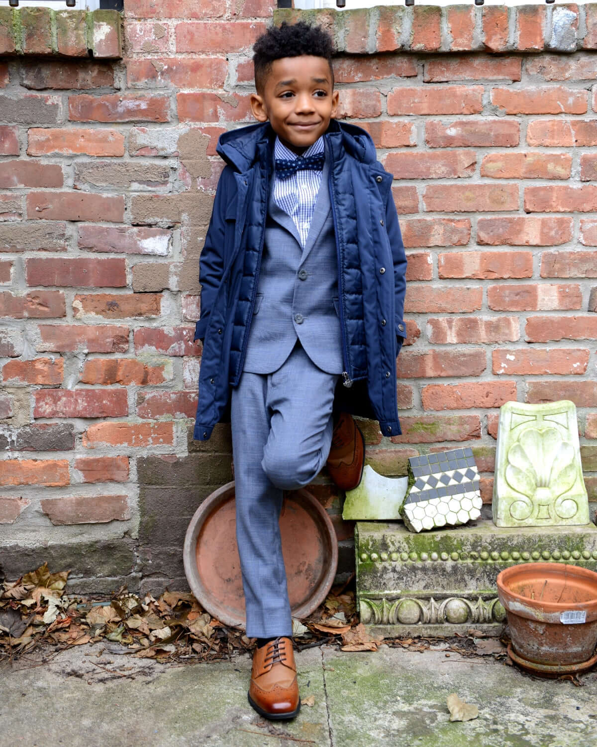 Little boy wearing Navy Blue Gotham Coat over a suit, styled for special events against a brick wall.