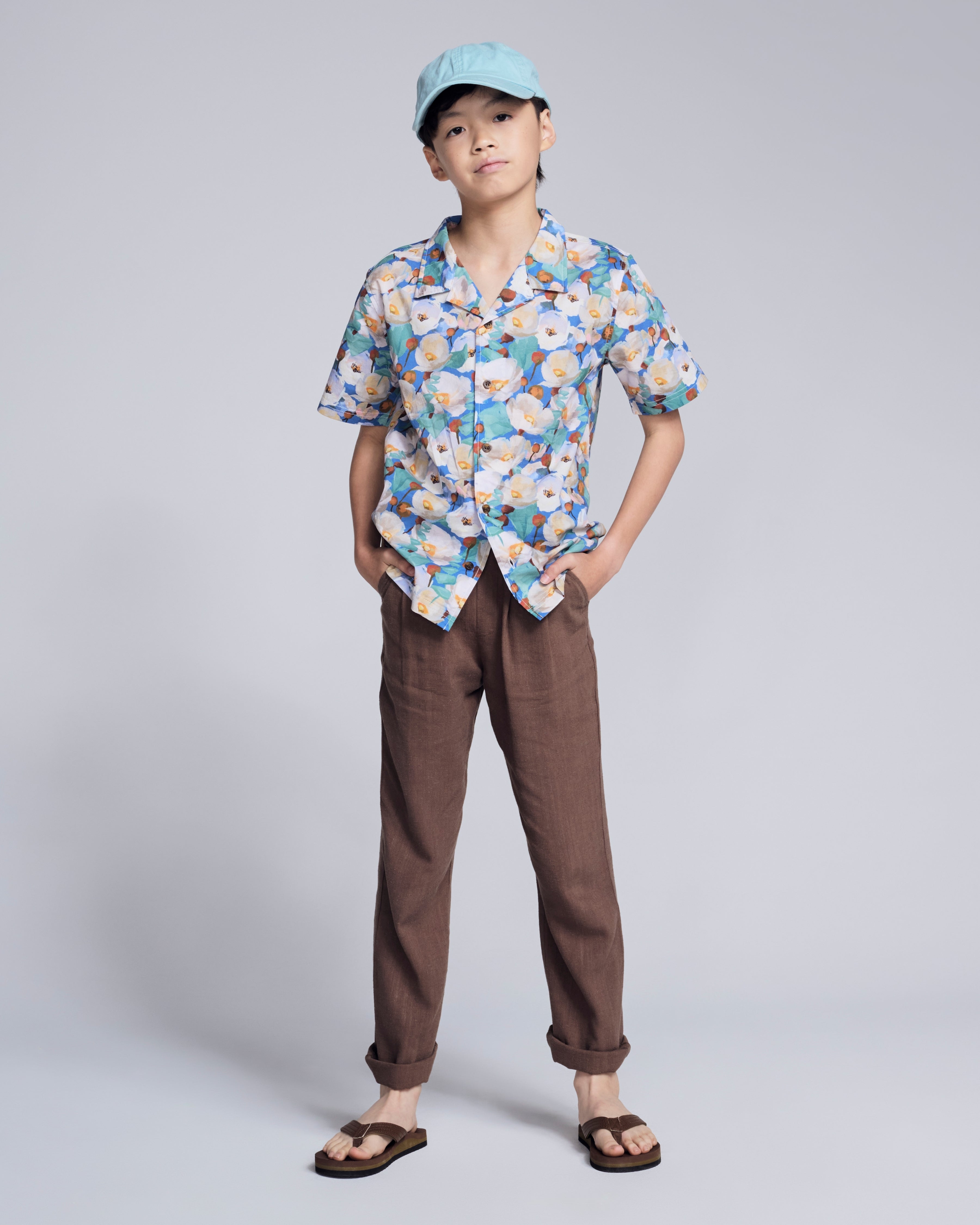 A boy wearing a Painters Garden Camp Collar Shirt with a floral pattern, paired with brown pants and a light blue cap.
