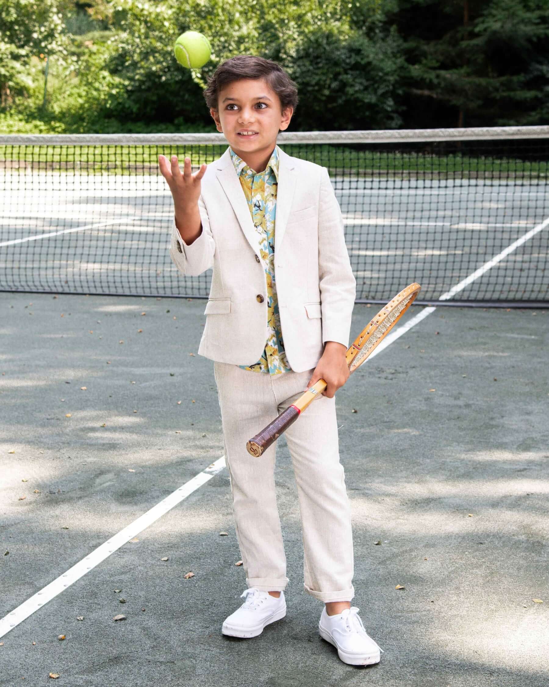 A young boy in a stylish linen blend Papyrus Sports Jacket, holding a tennis racket while juggling a tennis ball.