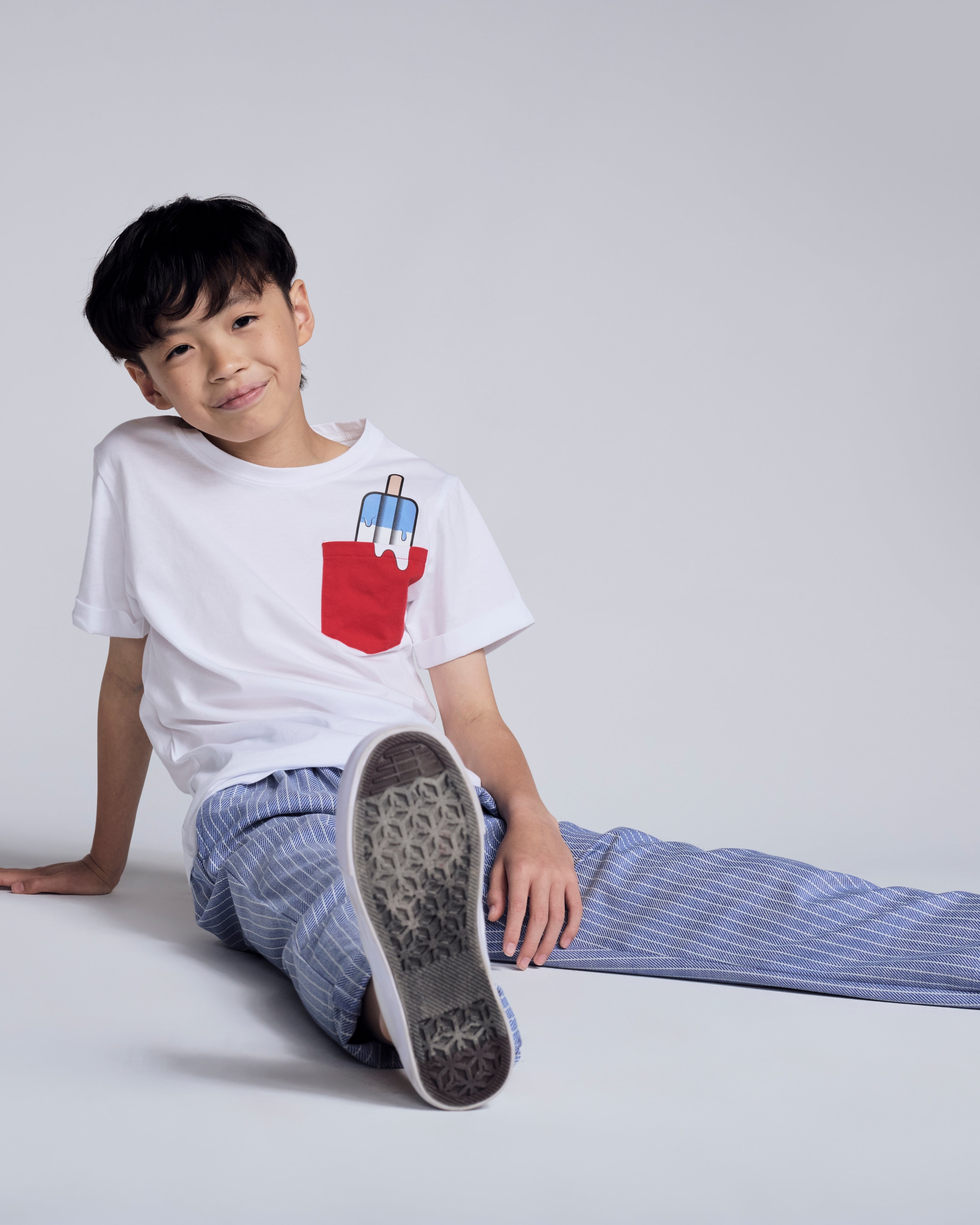 A young boy wearing the Popsicle White Day Trip Tee with a popsicle graphic pocket, sitting comfortably in casual attire.