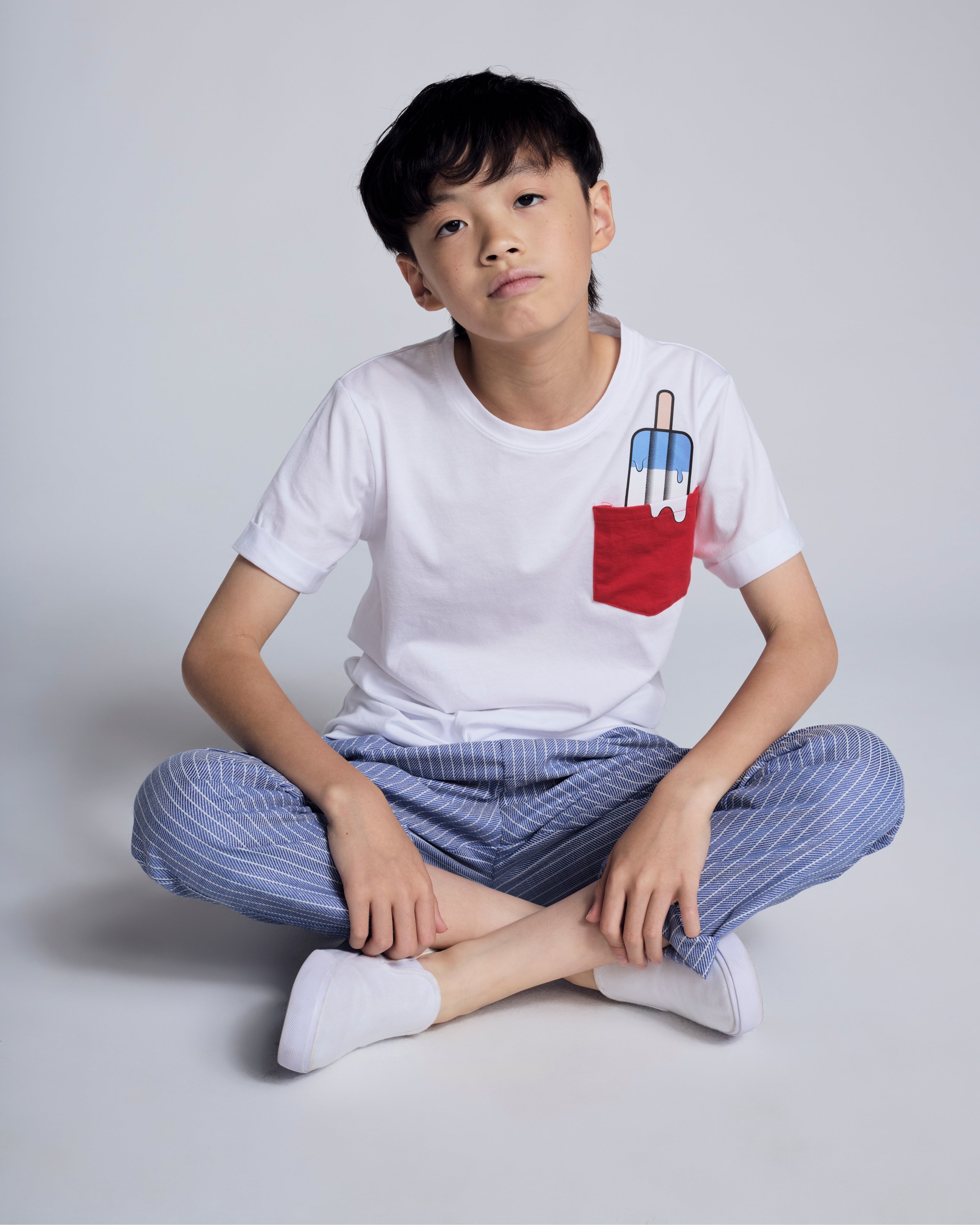 Boy sitting cross-legged wearing Popsicle White Day Trip Tee with popsicle graphic pocket.