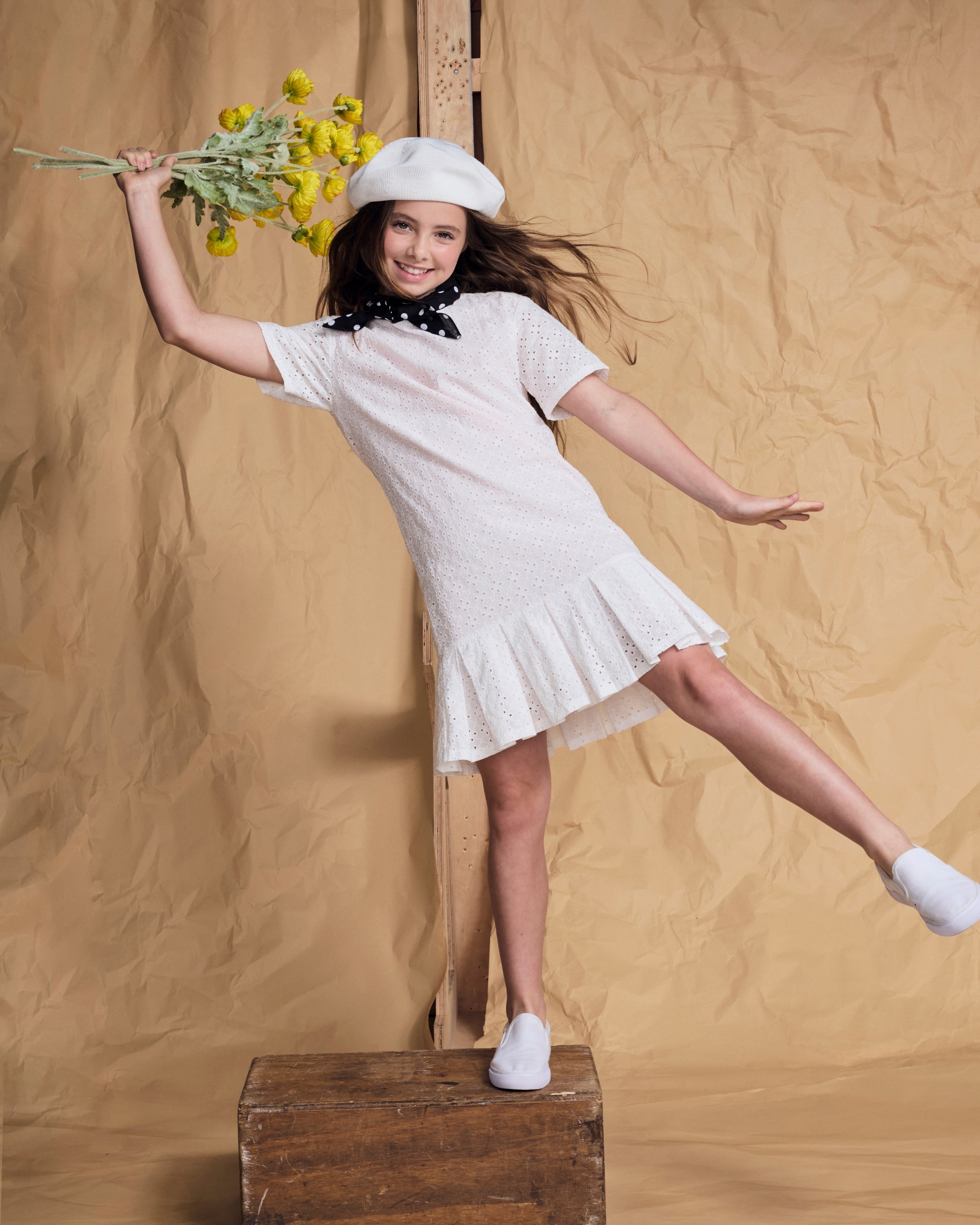 A girl twirling in a Soft Pink Eyelet Eve Dress, holding flowers, showcasing charm and playful style.