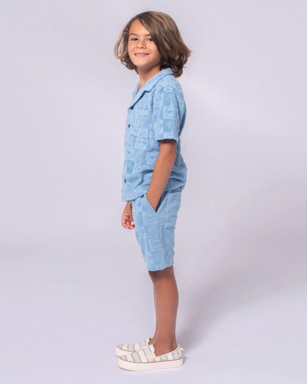 A child wearing Blue Resort Terry Shorts and Resort Shirt, showcasing playful palm tree and smiley face design.