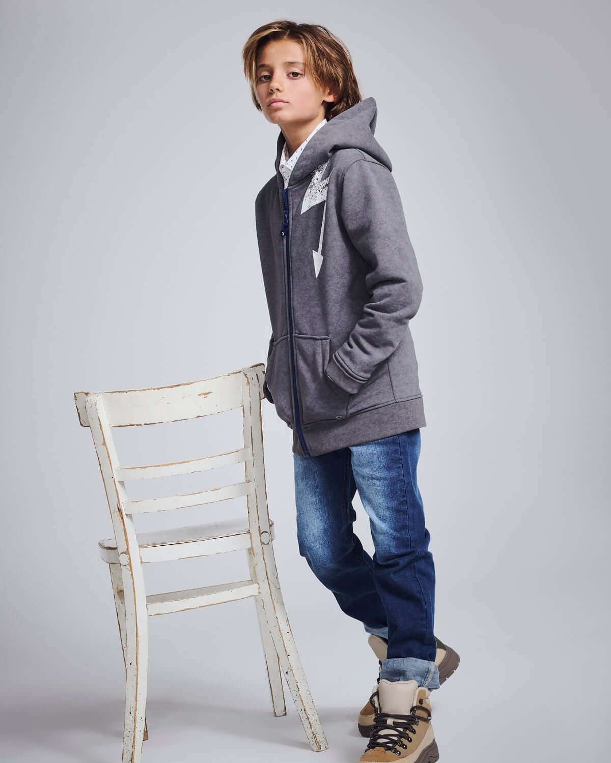 Boy in Medium Wash Slim Leg Denim and gray hoodie leaning on a chair, showcasing stylish kids' fashion.