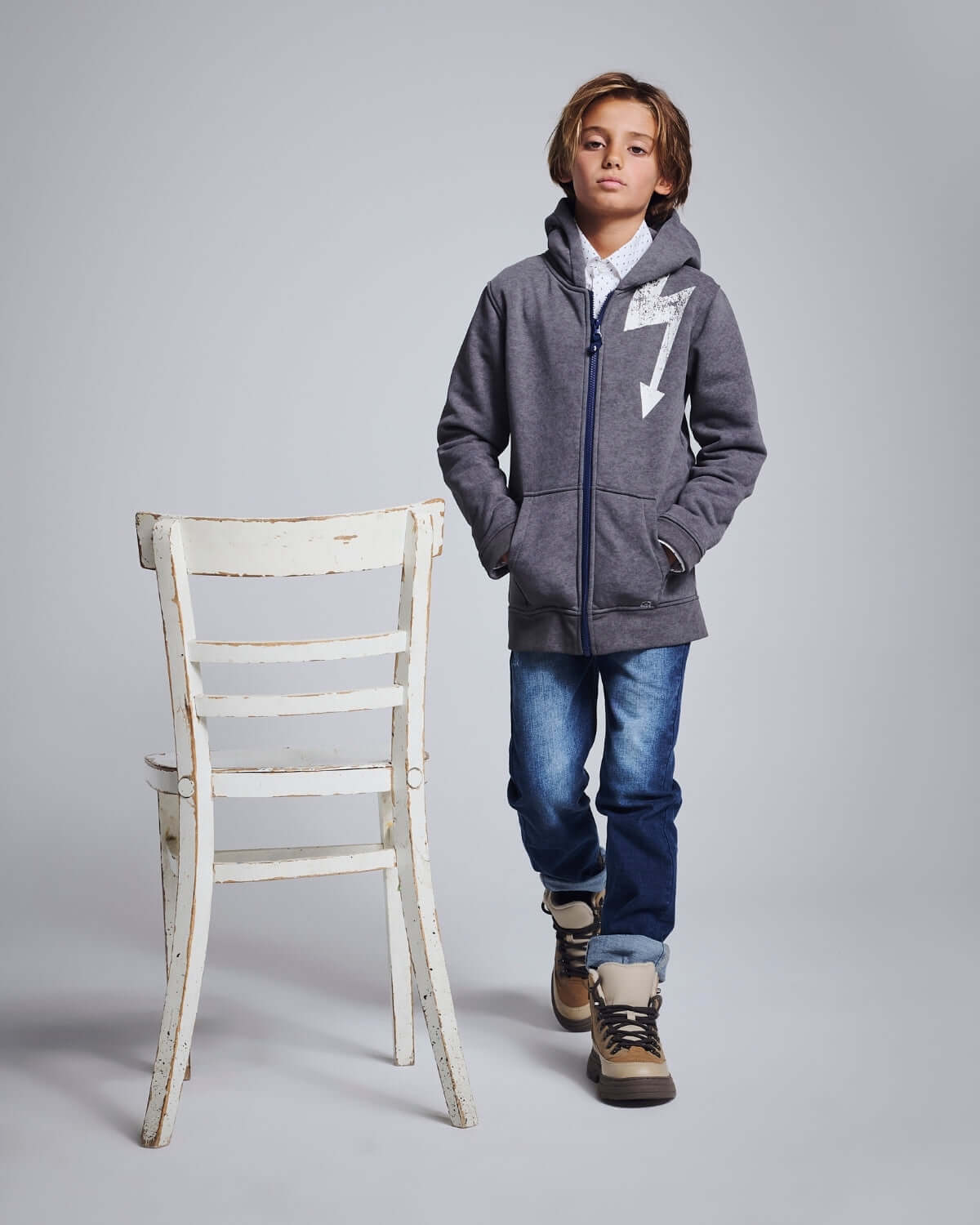 Boy wearing Medium Wash Slim Leg Denim with a grey hoodie, stylishly posing in front of a vintage chair.