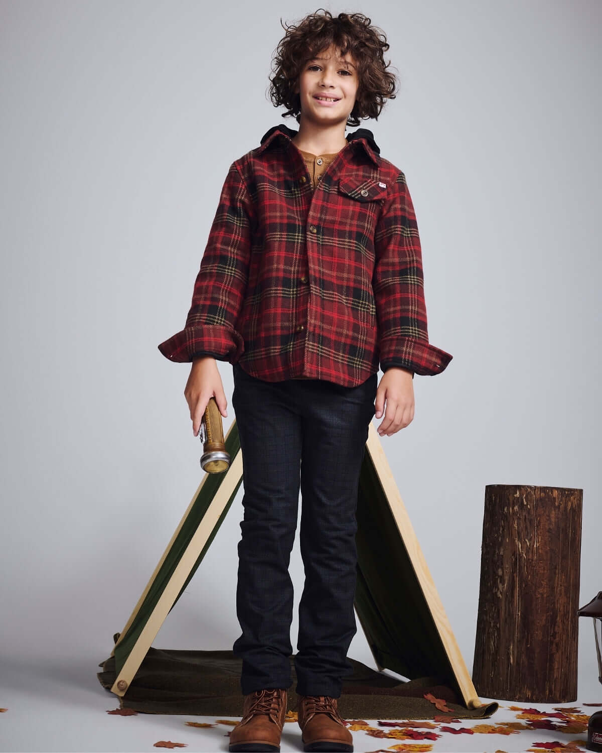 A young boy wearing a red plaid shirt and black jeans, standing in front of a tent with autumn leaves.