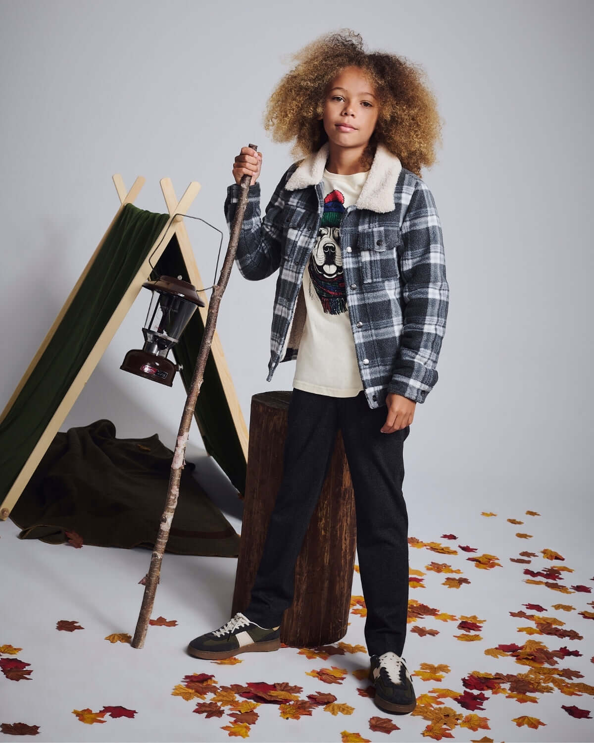 Kid wearing plaid shirt jacket and tweed everyday stretch pants, standing by a tent in autumn leaves.