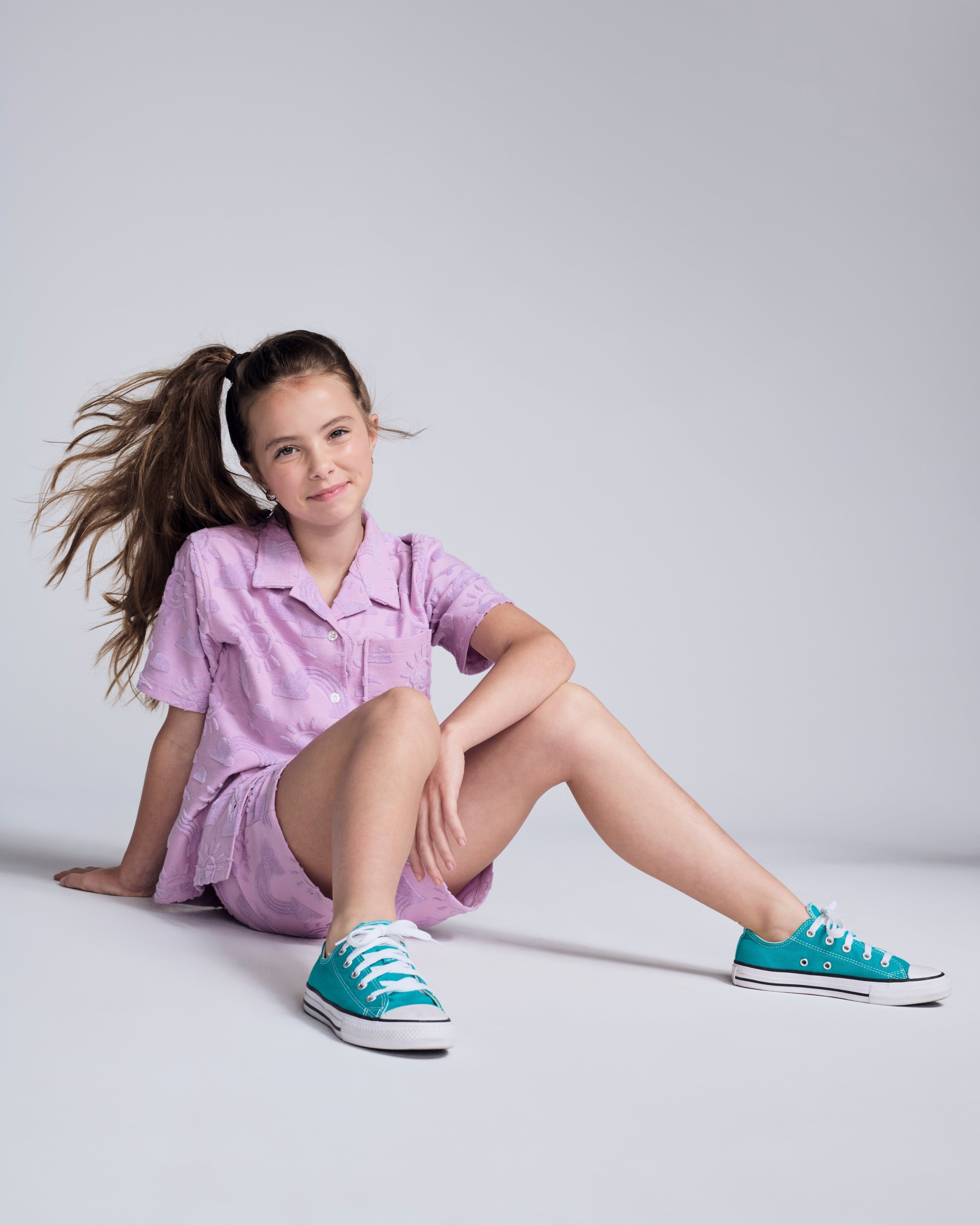 Girl wearing the Sunny Days Lilac Resort Shirt sitting and smiling with stylish sneakers.